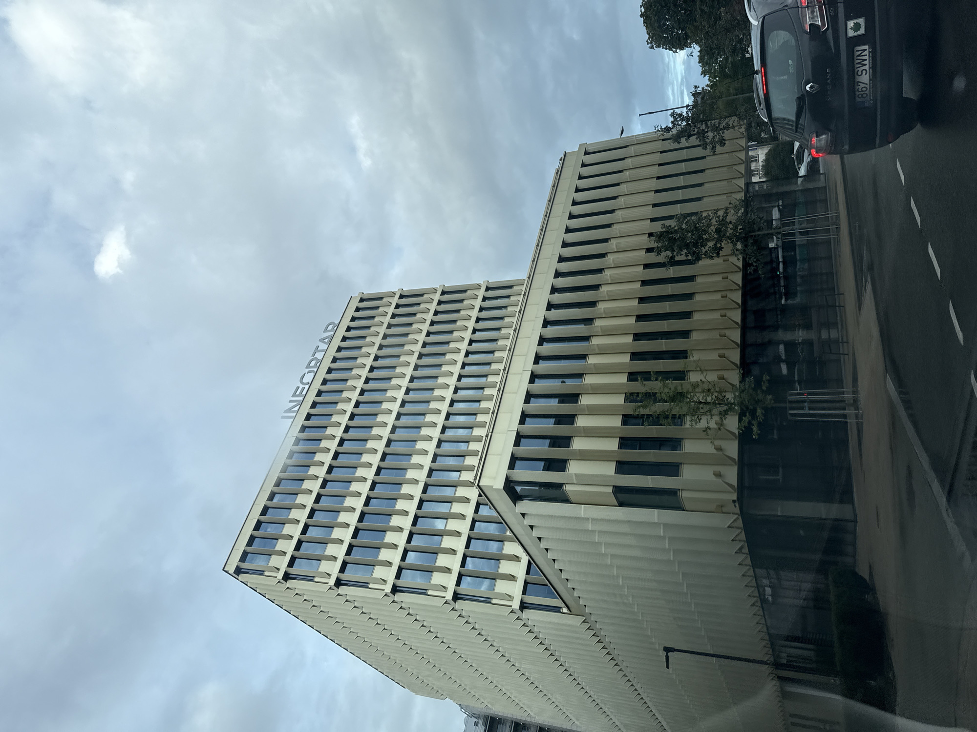 The image captures a modern office building with the signage "INEORTAR" visible at the top. The building features a repeating pattern of windows and vertical elements on its facade. In the foreground, a black car with license plate "867 SWN" is driving on a wet road, indicating recent rain. The setting appears to be an urban environment, consistent with Tallinn, Estonia, given the architectural style and general atmosphere. The sky is overcast with clouds.