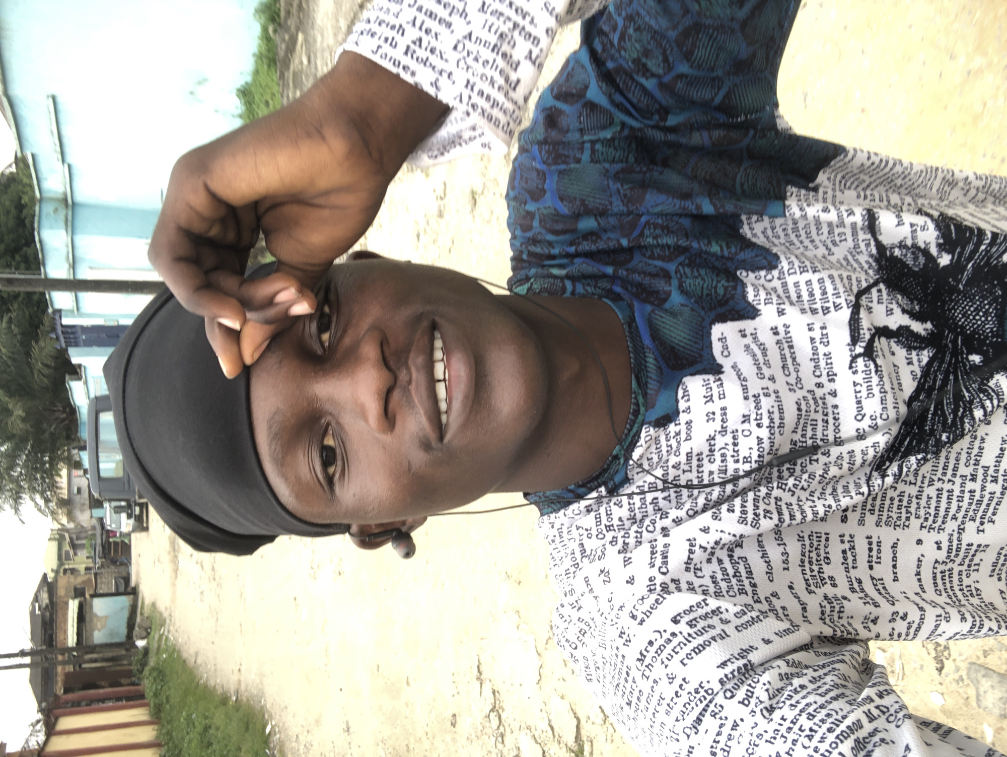 The image is a selfie taken outdoors on a dusty street in Ado, Nigeria. The subject, a young Black man wearing a black cap and a patterned shirt, is looking towards the camera, with his right hand touching his forehead. He has dark skin, dark eyes, and a faint smile. He is wearing earphones.
The background shows a street with houses and buildings on either side. The houses appear to be made of concrete and some have corrugated iron roofs. There are also trees and vegetation visible. Two vehicles, a black van and a smaller white vehicle, are parked on the street. The overall impression is of a residential or semi-commercial area within a city. The lighting suggests it is daytime. The ground is unpaved, consisting of dirt and gravel.