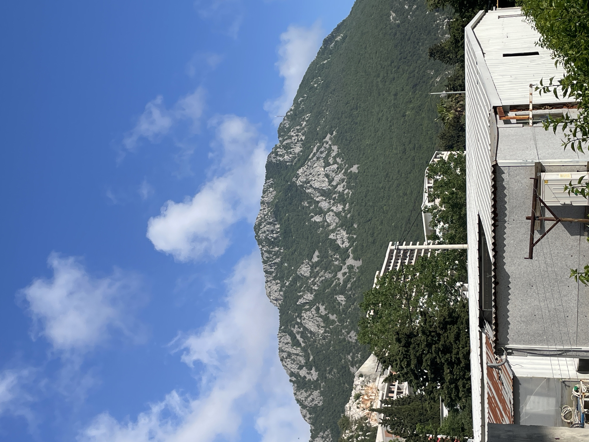 A vertically oriented, medium-shot photograph presents a mountain vista juxtaposed with residential structures. The left side of the image is dominated by a clear blue sky dotted with fluffy white clouds. Adjacent to the sky, a large mountain covered in dense green vegetation rises prominently, its rocky terrain interspersed with patches of grey rock.
To the right of the mountain, buildings with light-colored walls and corrugated roofs partially obstruct the view. Trees with dense foliage are interspersed between the buildings. The photo exhibits a slightly tilted perspective. The location is not explicitly identified as Čanj, Montenegro, based solely on visual evidence.