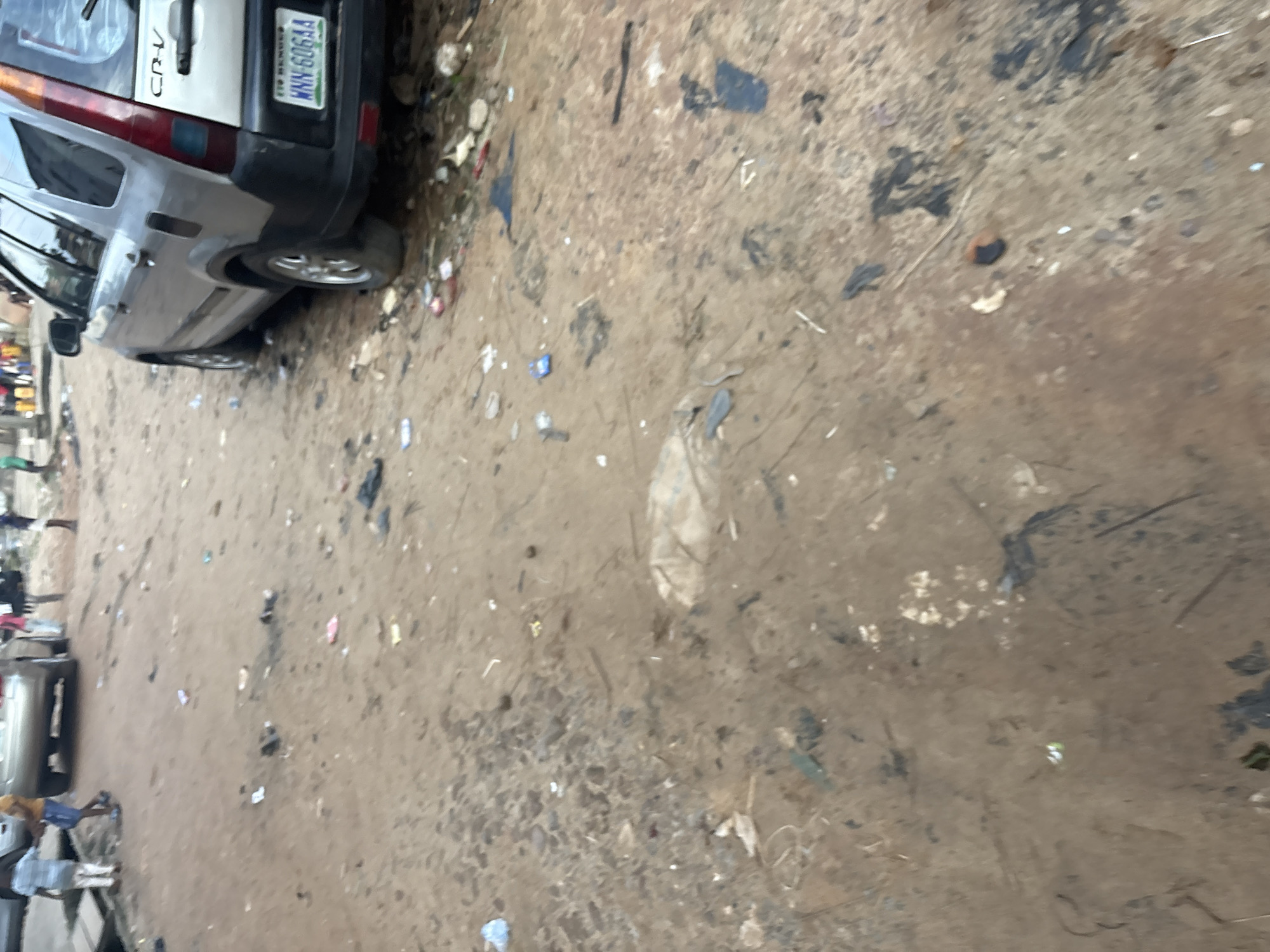 The image depicts a street scene in Onitsha, Nigeria, characterized by a dirt road strewn with litter. In the foreground, the ground is a mixture of dirt and debris, including small pieces of plastic and other refuse. A few children are visible in the background, some standing near a vehicle. One child in a blue and white striped shirt and white pants is holding hands with another child wearing a yellow shirt and blue shorts. Further down the road, a silver Honda CR-V SUV with license plate WNN 606AA is parked. The overall environment appears unkempt and indicative of a densely populated urban area.