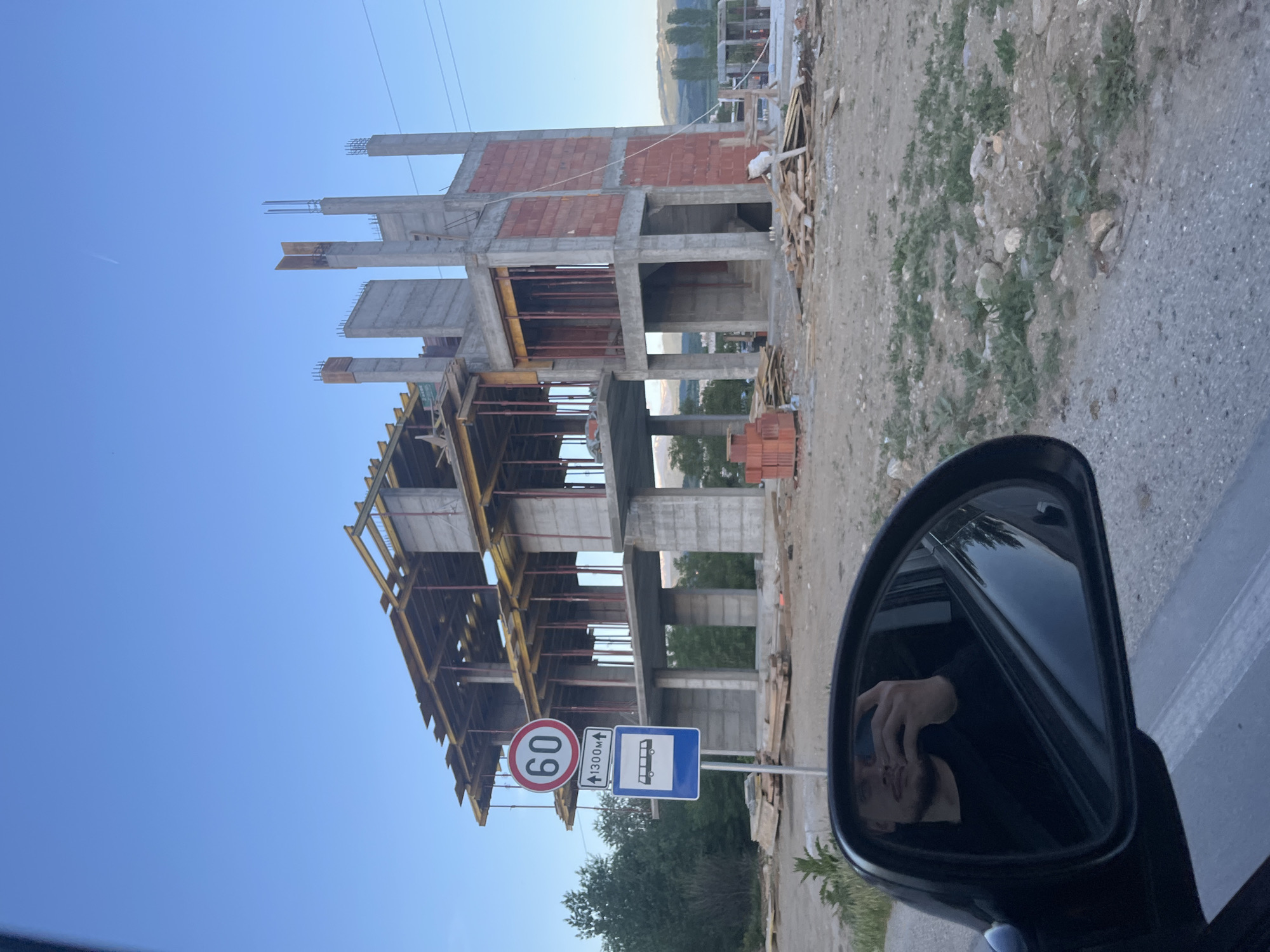 The image shows an unfinished building under construction. The building has a concrete frame with some brick walls. There is wooden scaffolding around the lower floors. A speed limit sign (60) and a bus stop sign are visible in the foreground. In the car's side mirror, a man is reflected taking a photo. Based on the presence of a bus stop with Cyrillic script and additional information, the location is likely near a construction site in Blagoevgrad, Bulgaria. The surrounding area appears to be semi-urban with a mix of construction and natural vegetation.
