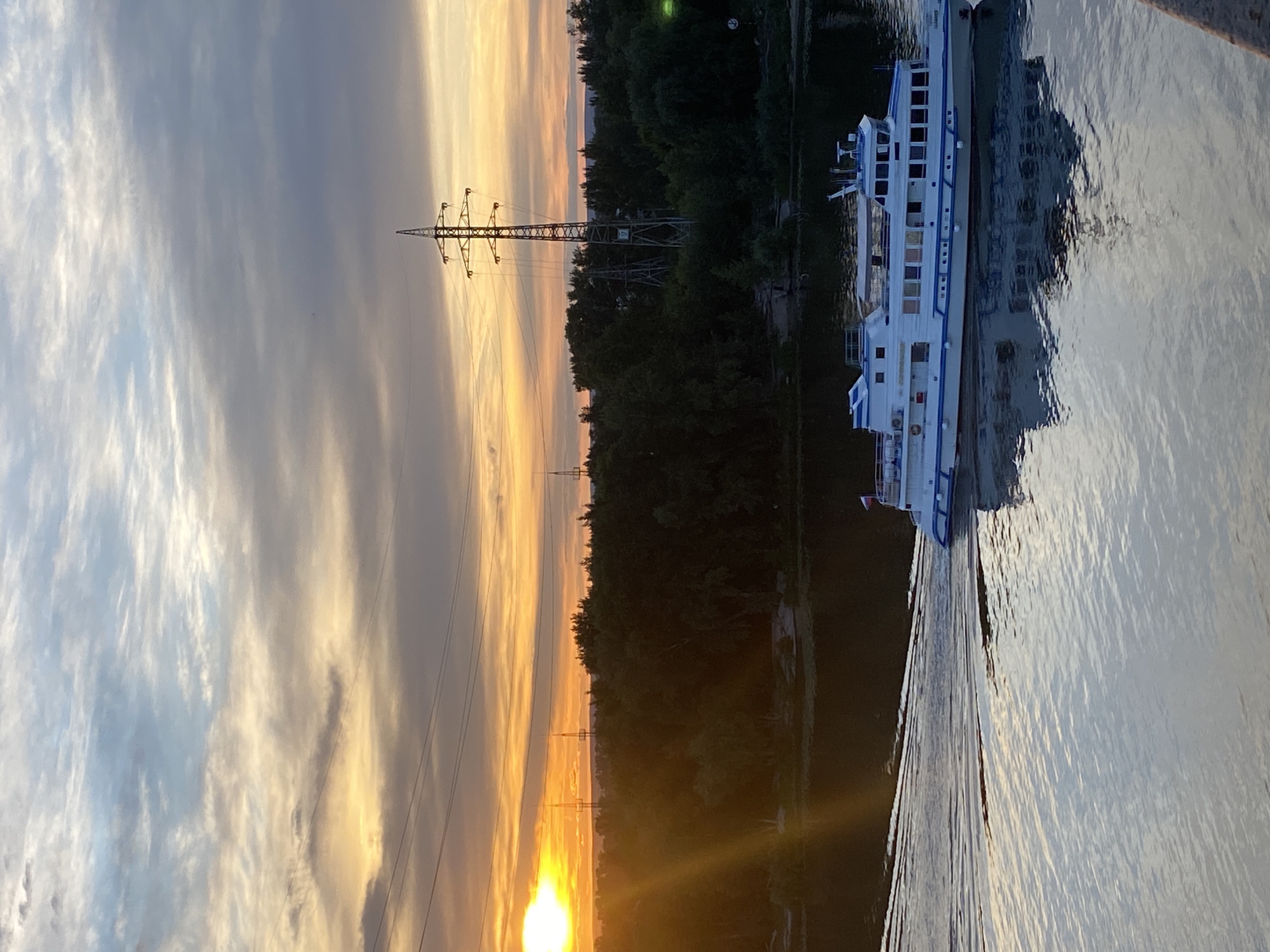 An evening sunset casts a warm glow over a wide river in Novokuybyshevsk, Russia. The sky is a gradient of oranges, yellows, and grays, with scattered clouds. In the distance, a dense line of trees forms the horizon, punctuated by tall, skeletal power line towers. A white passenger boat with blue trim moves from right to left across the frame, creating ripples in the water. The boat is reflected in the calm surface of the river. The image is taken from a slightly elevated perspective, suggesting the viewer might be on a bridge or a higher vantage point.
