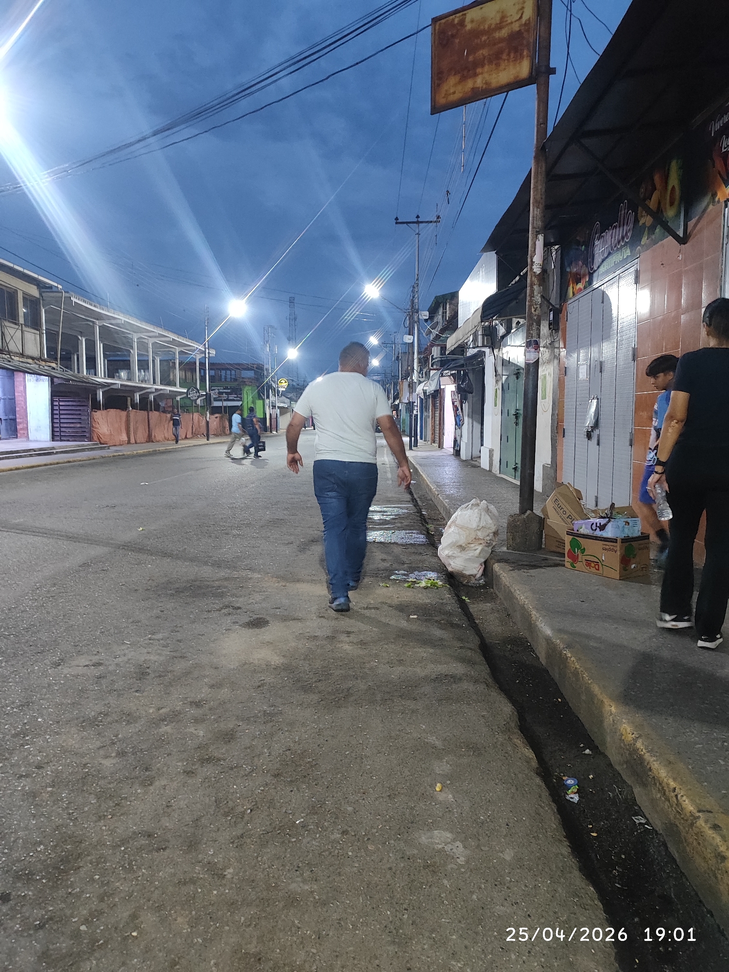 The image captures an urban scene in Tinaquillo, Venezuela, at twilight, as indicated by the date stamp in the lower right corner: 25/04/2026 19:01. A man wearing a white t-shirt and jeans walks away from the camera down the center of a paved road. Several people can be seen in the distance further down the street. To the right of the frame, two individuals stand on the sidewalk near closed storefronts, one in black and the other with a blue shirt. A white plastic bag and cardboard boxes lie on the sidewalk next to them. Overhead streetlights illuminate the road, and utility lines stretch across the sky. The buildings lining the street appear to be commercial establishments, and a rusty signpost stands on the sidewalk. The street surface is worn, with signs of age and use. A gutter runs alongside the sidewalk.