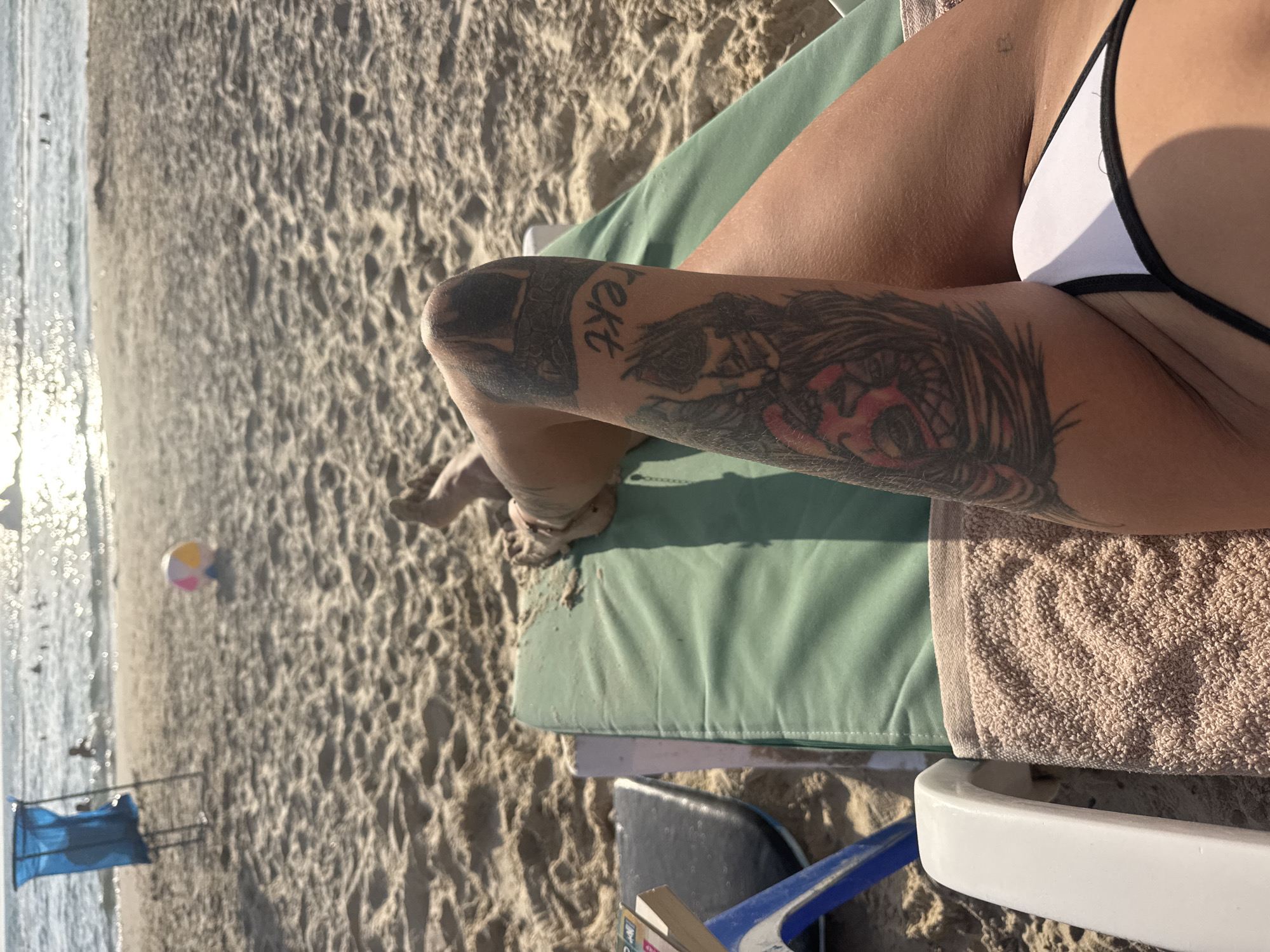 The image shows a close-up view of a person relaxing on a beach lounge chair. The person is wearing a white bikini top. Their left arm, adorned with a large, intricate tattoo, rests on a folded towel. The tattoo appears to depict a stylized eye and other dark imagery. Part of the person's leg is also visible, with sand clinging to their skin.
The setting is a sandy beach with the ocean in the background. Sunlight glints off the water, suggesting it is daytime, likely late afternoon given the warm, angled light. The sand has visible ripples and texture, indicating a dry beach. A colorful beach ball, a blue beach tent, and what appear to be people in the distance suggest a recreational beach environment. The overall mood is relaxed and leisurely, typical of a beach vacation. There is some visible text on the tattoo, though it is partially obscured and difficult to read clearly.