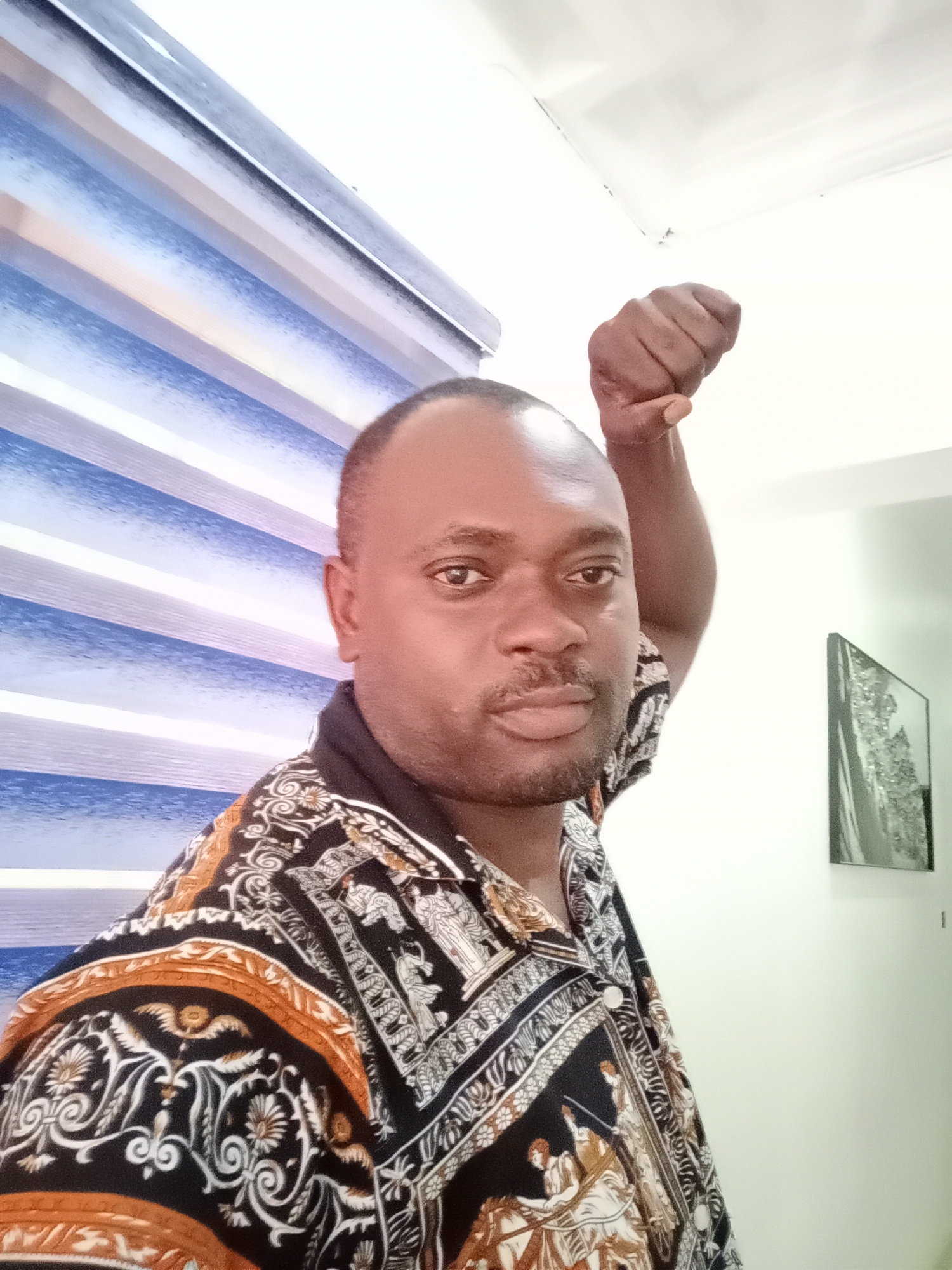 A medium shot depicts a Black man with a shaved head and a short beard. He's wearing a patterned short-sleeved shirt with a dark collar and is posing with his fist raised in the air. The background features horizontal blue and white blinds to the left, and a white wall with a framed picture on the right. The location appears to be inside a building in Gwarinpa, Nigeria.