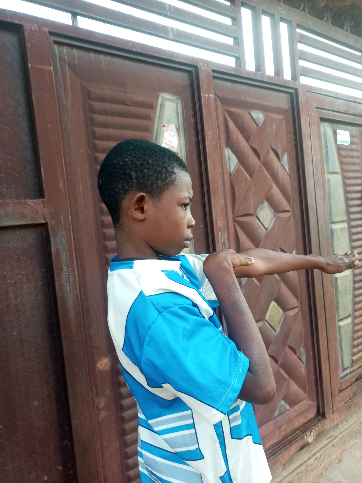 A young boy stands in front of a reddish-brown metal door with a geometric pattern. He is wearing a blue and white t-shirt with horizontal stripes on the sleeves and lower half. His right arm is extended outwards, with his hand open and palm facing forward, as if to signal or stop someone. The boy's left arm is bent at the elbow, with his hand resting on his upper arm. The background appears to be an outdoor setting, possibly in the city of Tempure, Nigeria, with a textured wall or pavement visible at the bottom of the frame. The lighting suggests it is daytime, and the overall impression is a candid shot.