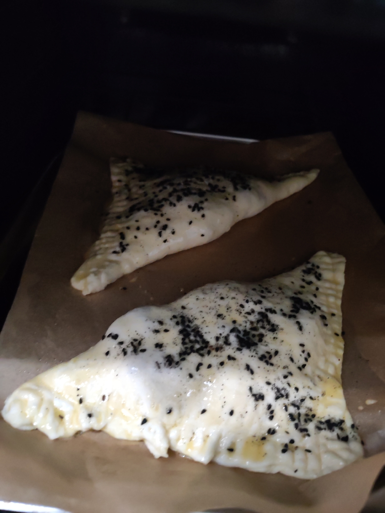 The image displays two uncooked pastries, possibly samosas, samsa, or similar hand pies, placed on a sheet of brown parchment paper. They are positioned on what appears to be a baking tray inside a dark, enclosed space, likely an oven. The pastries are roughly triangular in shape with visibly crimped edges, suggesting they are sealed. Both are coated with a light, shiny glaze, characteristic of an egg wash, and generously sprinkled with black sesame seeds across their top surfaces. The dough appears raw or lightly proofed, indicating they are either ready to be baked or are in the initial stages of baking. The background is dark and out of focus. There are no people, discernible text, or specific environmental cues to determine the time of day, weather, or the precise location within Izhevsk, Russia.