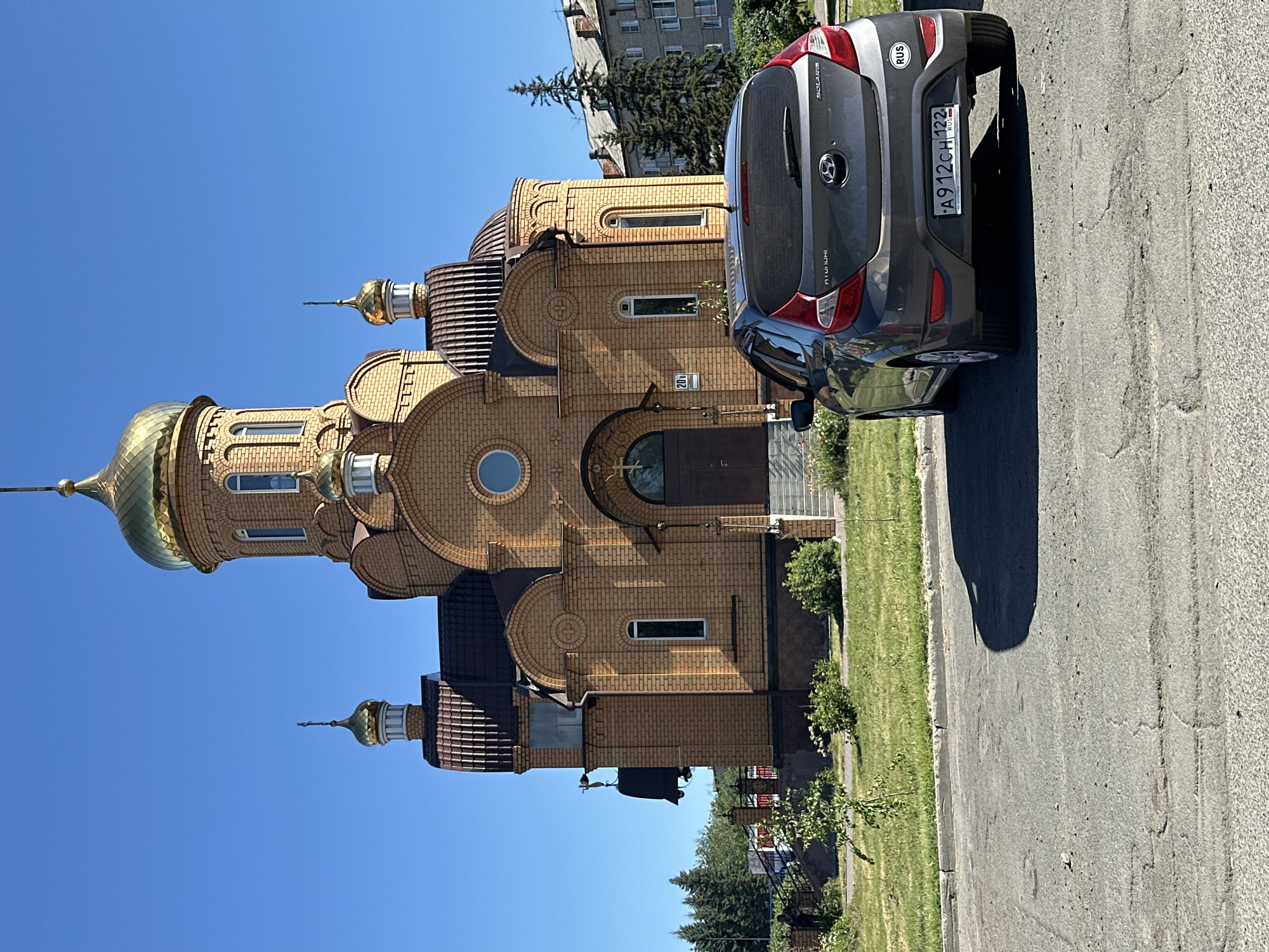 A dark gray Hyundai Solaris car is parked on a cracked asphalt road in front of a tan brick Orthodox church with gold domes. The church has a tall central dome and several smaller domes, as well as arched windows and entrances. The car's rear license plate reads "A912CH 122". A small circular "RUS" sticker is visible on the right side of the car's bumper. The church is situated in an area with green grass and some landscaping. In the background, there are evergreen trees and a multi-story residential building. The sky is clear and blue. The scene appears to be in Novoaltaysk, Russia.