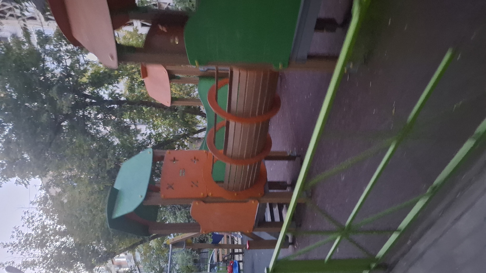 The image captures a section of a playground in Shymkent, Kazakhstan, featuring a train-themed play structure. The structure, primarily made of wood and brightly colored plastic, includes sections painted orange and green, resembling train cars. A cylindrical tunnel, designed to look like a train car, is visible in the center, with orange rings at either end. The ground of the playground appears to be a soft, dark surface, likely rubberized for safety. A green metal fence runs across the foreground, partially obscuring the view of the playground equipment. Trees with green foliage are visible in the background, along with apartment buildings. The sky is overcast, suggesting cloudy weather. No people are present in the image.