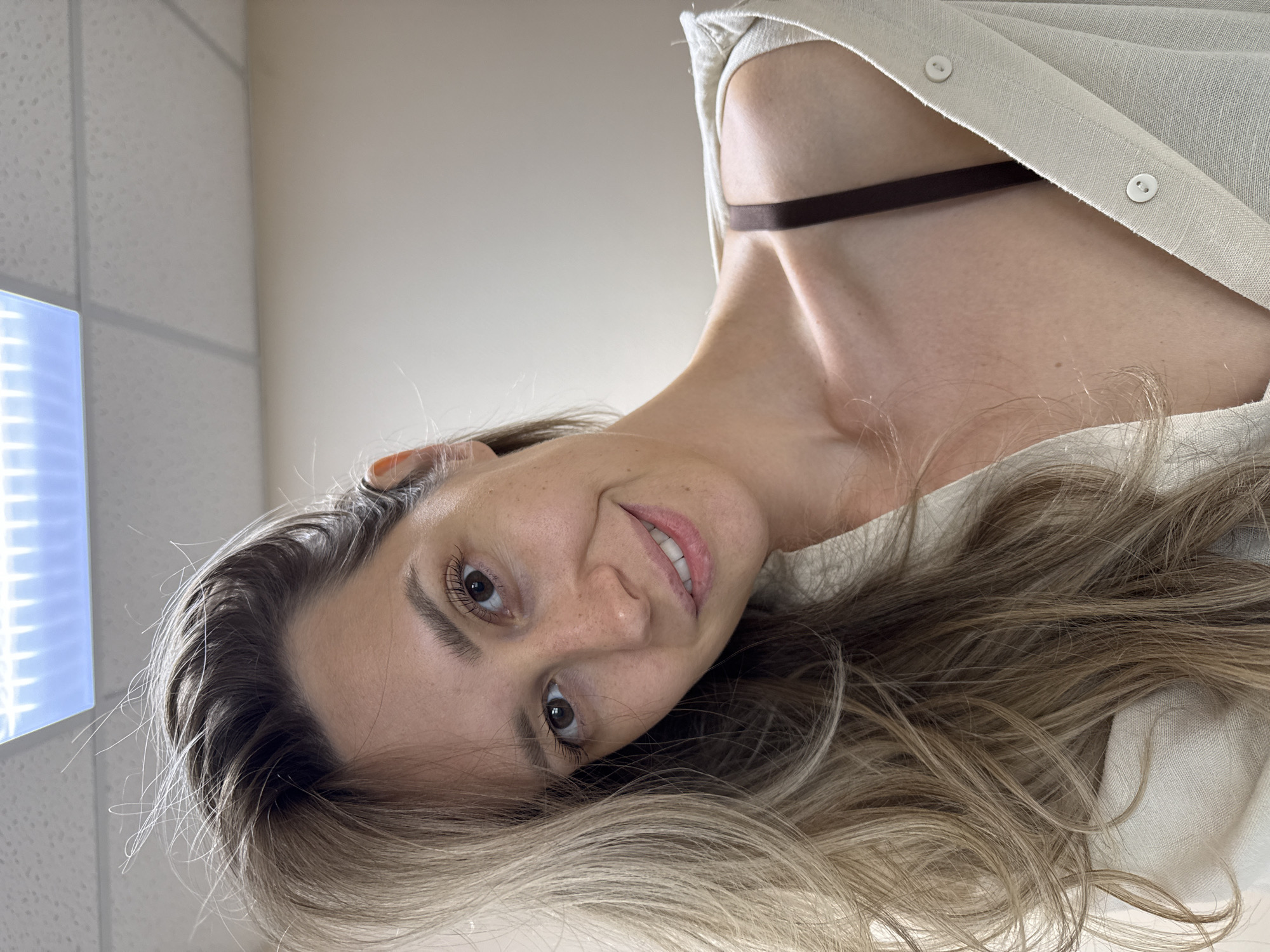 A young woman with long, blonde hair is taking a selfie in an indoor setting. She is wearing a light-colored button-up shirt, partially open to reveal a brown bra strap. The background appears to be an office or similar commercial space, with a fluorescent light fixture visible on the ceiling. The woman is smiling and looking directly at the camera. There are no other people or significant objects in the frame.
