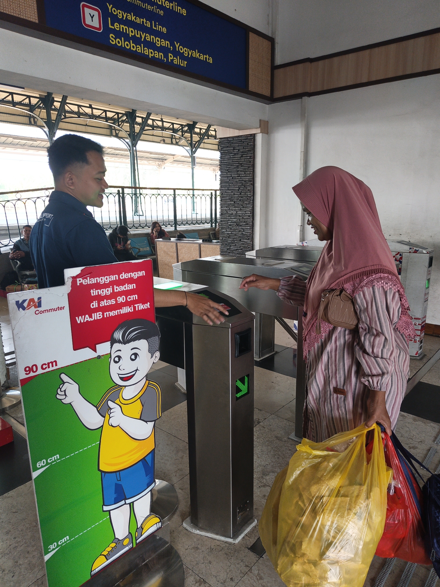 An Indonesian man in a dark blue uniform stands at an automated ticket gate, looking to his left. He is wearing a dark blue polo shirt with "SPORT" visible on the sleeve. To his right, a woman wearing a pink hijab and a striped long-sleeved shirt is interacting with the ticket gate. She is holding several shopping bags. In the foreground, a large red and green sign displays a cartoon boy and a height chart with markings at 30 cm, 60 cm, and 90 cm, along with Indonesian text. The background shows a covered train platform with people sitting on benches and another ticket gate. The scene is likely at a train station in Klaten, Indonesia.
