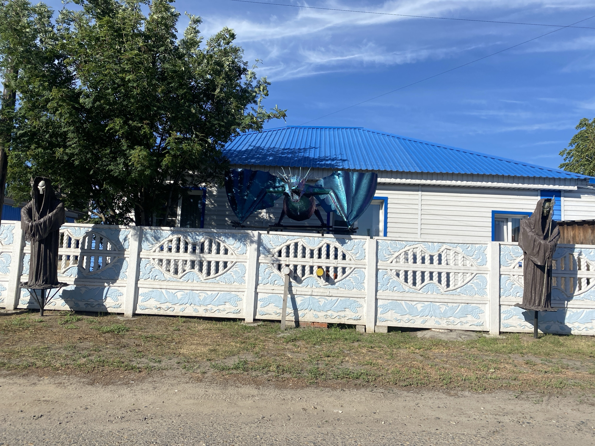 In Mamontovo, Russia, a white house with a bright blue metal roof is visible behind a decorative fence. Two figures, resembling the Grim Reaper, stand on either side of the property, flanking the entrance. One figure is partially obscured by a large green tree to the left. A large, metallic blue dragon sculpture is prominently displayed on the house's facade, positioned between two windows. The fence itself is made of concrete panels, adorned with white ornate patterns and light blue vine motifs. The ground in front of the fence is unpaved, with patches of dry grass and dirt. The sky above is a clear blue with wispy white clouds.