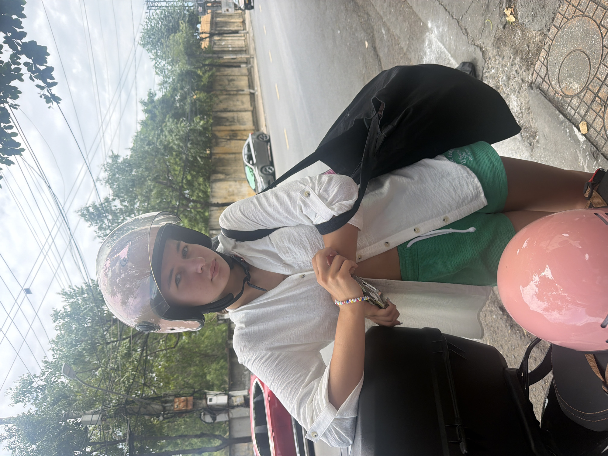 A young woman, wearing a white button-down shirt and green shorts, poses with a scooter in Nha Trang, Vietnam. She has a helmet on her head and a black bag slung over her shoulder. The scooter has a black storage box and a pink helmet rests on the seat. The background shows a street scene with trees, a wall, and cars. The sky is cloudy.
