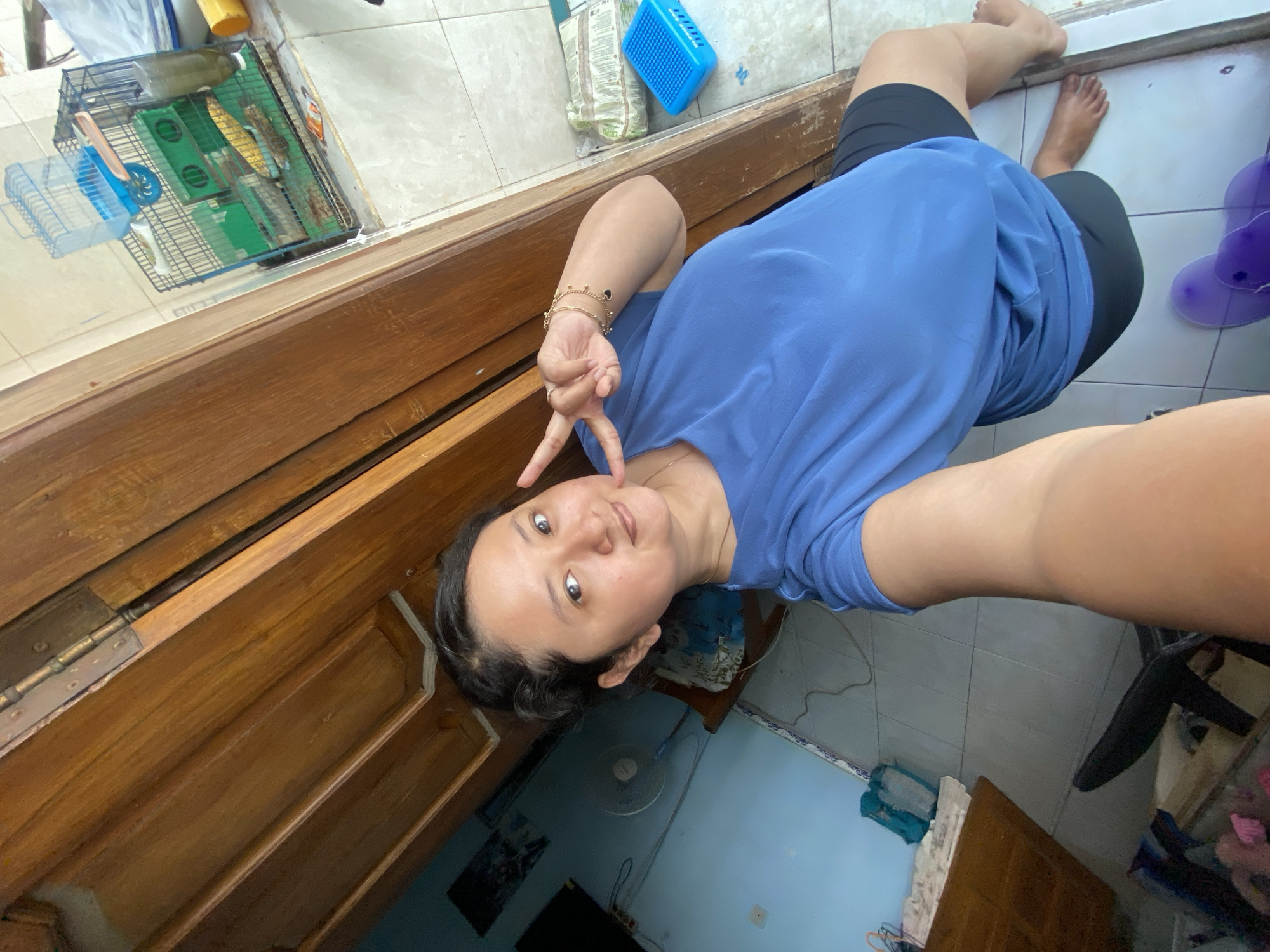 The image captures a casual selfie taken by a woman indoors, likely in a home setting in Gelam, Indonesia, during the daytime. The woman, appearing relaxed and calm, is positioned lying down, possibly on a wooden bed frame or bench, looking up towards the camera. She is wearing a blue t-shirt and black shorts or leggings, and is barefoot. Her left arm is extended, presumably holding the camera, while her right hand is raised near her face, forming a peace sign. She also wears a thin gold bracelet on her left wrist.
The background reveals various elements of a domestic space. The floor is covered with light-colored tiles. In the upper left portion of the image, a wire pet cage is visible, containing a water bottle, a feeding dish, and bedding, suggesting the presence of a small pet like a hamster. Next to the cage are a blue plastic container and a bag with partially legible text that appears to say "CHUP...", possibly from a "Chupa Chups" product. The lower right background features a blue-painted wall and a floor fan. Other items, such as miscellaneous clothes or fabrics, are casually placed on the floor or furniture, contributing to a lived-in atmosphere. A robust wooden structure, possibly part of the furniture the woman is resting on, occupies the left side of the frame, showing a metal hinge. The lighting is bright and even, consistent with daylight.