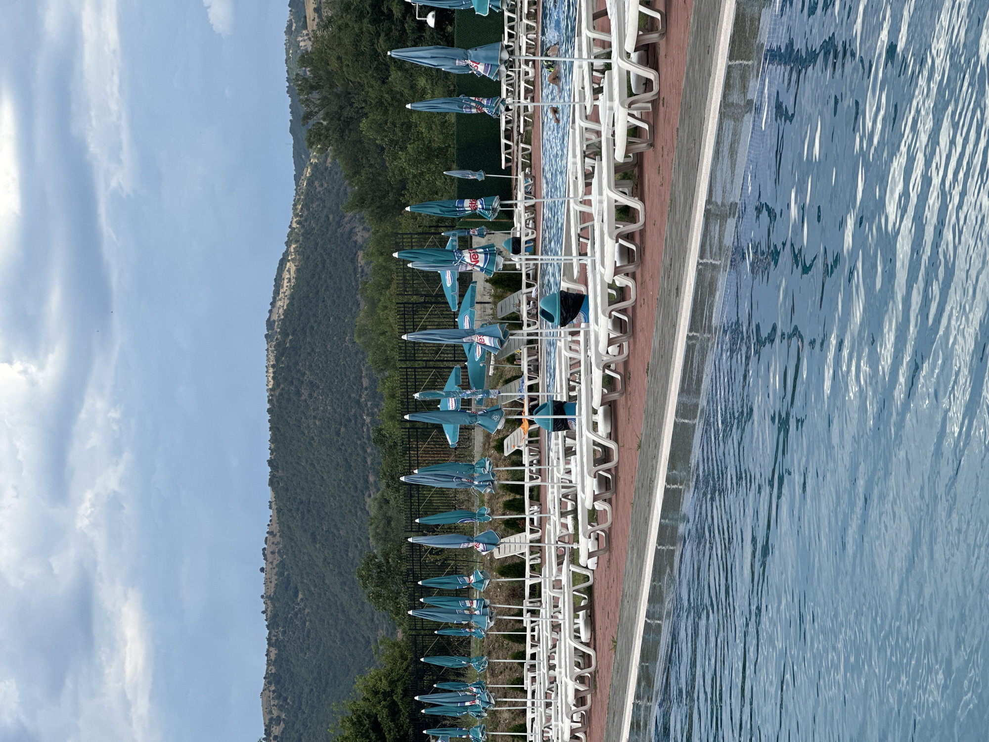 The image depicts a poolside scene with numerous closed turquoise umbrellas lined up next to a swimming pool. White lounge chairs are visible behind the umbrellas, and a person is partially submerged in the pool water. The pool has a tiled edge and the water is a clear blue with ripples reflecting the sunlight. In the background, a lush green mountainside rises steeply, covered in trees. The sky above is a pale blue with scattered white clouds. The overall impression is of a tranquil, sunny day at a resort or recreational area. The scene is likely set in Simitli, Bulgaria.