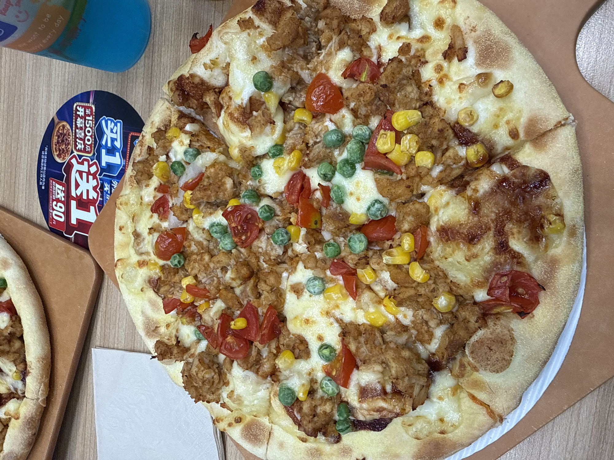 A close-up, overhead shot captures a whole pizza, with its crust visible at the bottom and right edges, resting on a light brown surface, possibly a pizza peel or cutting board. The pizza is topped with shredded chicken, corn kernels, peas, and chopped cherry tomatoes, all covered in melted cheese. To the left of the pizza, a white napkin is partially visible, and to the upper left, a sliver of another pizza is seen. In the upper right corner, a blue plastic cup with a black lid is present, along with a circular promotional flyer with Chinese text and images of pizza. The background is a wooden table surface. The scene is located in Dongcheng District, China.