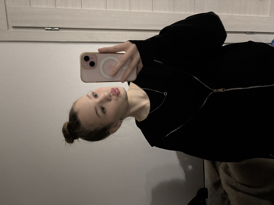 A selfie of a young woman with her hair in a bun, pursing her lips playfully. She's wearing a black, zip-up hoodie and a delicate silver necklace. Her phone, with a light pink case and a magnetic ring on the back, is held up to capture the image. The background features a neutral-colored wall and what appears to be a textured door or cabinet. A hint of a cozy, soft blanket is visible at the bottom right.