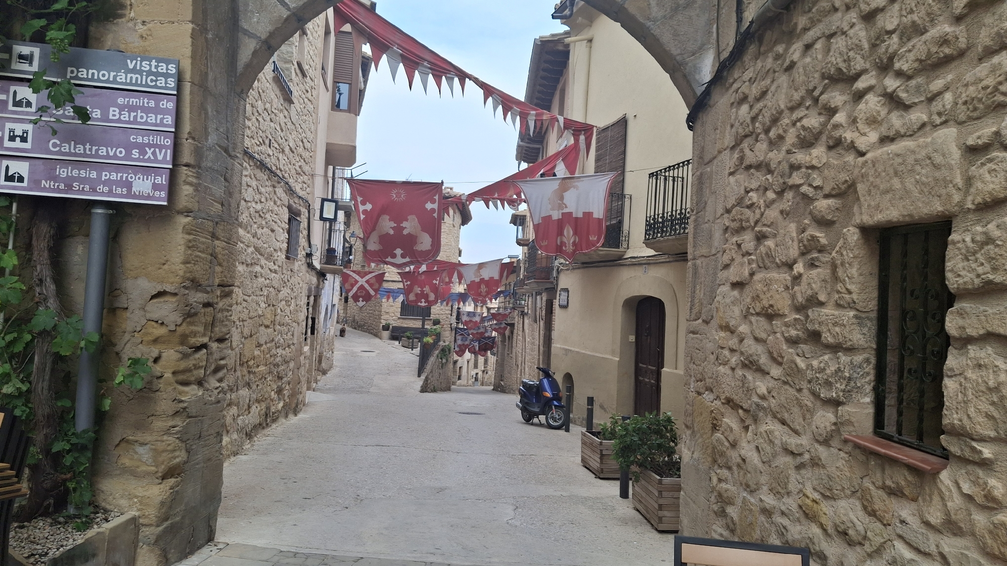 The image shows a narrow, stone-paved street in La Fresneda, Spain, adorned with red and white banners and pennants, likely for a festival or historical reenactment. The street is flanked by old stone buildings with wooden shutters and wrought-iron balconies. A blue scooter is parked on the right side of the street. A signpost on the left indicates directions to "vistas panorámicas" (panoramic views), "ermita de Santa Bárbara" (hermitage of Saint Barbara), "castillo Calatravo s.XVI" (Calatrava castle, 16th century), and "iglesia parroquial Ntra. Sra. de las Nieves" (parish church Our Lady of the Snows). The architecture and signage suggest a historic town, possibly with medieval origins. The sky is overcast.