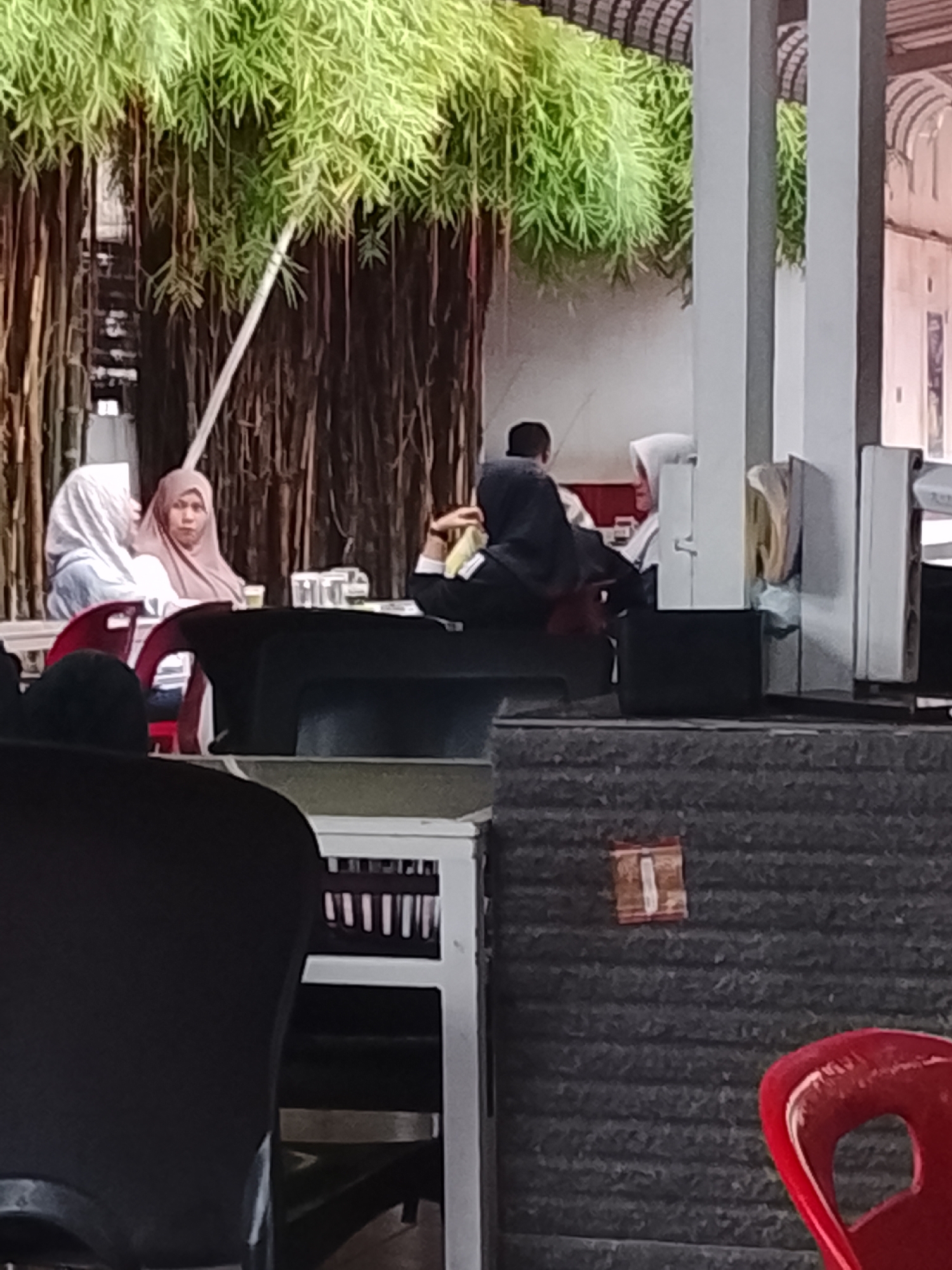 The image captures a group of people sitting at tables, seemingly in an outdoor dining area, in Lhokseumawe, Indonesia. A woman wearing a tan hijab is looking directly at the camera. Other women with hijabs are also present. There's foliage overhead, possibly from trees or vines, and architectural elements like pillars or walls in the background. Tables and chairs are visible in the foreground and midground.