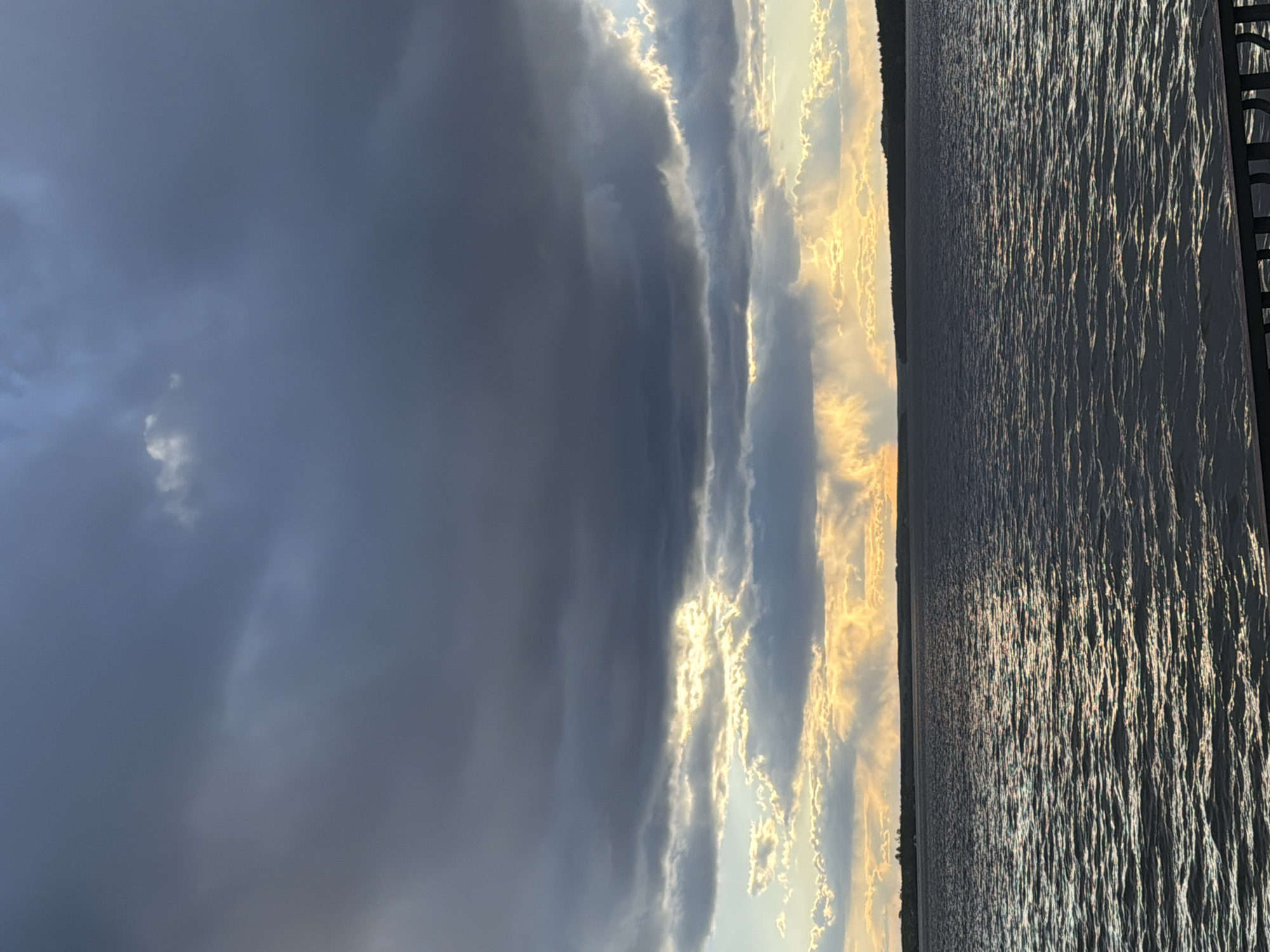 The image depicts a view of a body of water, possibly a lake or river, at sunset in Votkinsk, Russia. The sky is divided between dark, stormy clouds on the left and illuminated, golden-edged clouds on the right. The water reflects the sunlight, creating a shimmering effect. A shoreline is visible along the horizon. A portion of a wooden structure, possibly a dock or pier, is visible on the right.