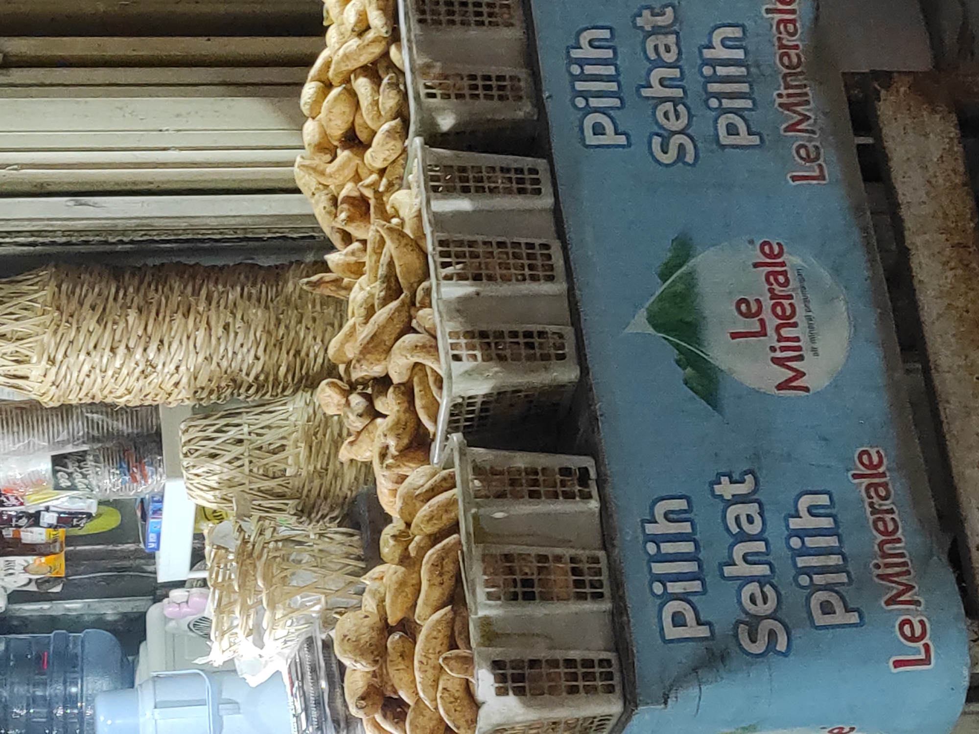 The image depicts an outdoor market stall, likely in Panenjolan, Indonesia, showcasing a display of sweet potatoes and bottled water. In the foreground, three white plastic baskets filled with sweet potatoes are arranged on a light blue surface. The blue surface features text in white and red. The white text reads "Pilih Sehat Pilih" which translates to "Choose Healthy Choose". Below this, in red, is "Le Minerale", the brand name of bottled water. To the left of the text, a logo for Le Minerale is visible, featuring a water droplet shape with mountains inside.
Behind the sweet potatoes, several woven baskets of various sizes are stacked. The stall appears to be illuminated by artificial light, creating a somewhat warm and slightly dim atmosphere. In the background, there are glimpses of other market elements, including plastic containers, packaging, and possibly more woven goods. No people are clearly visible in the frame, suggesting the stall is either unattended or the vendor is out of view. The overall impression is a functional retail setting with fresh produce and beverages for sale.