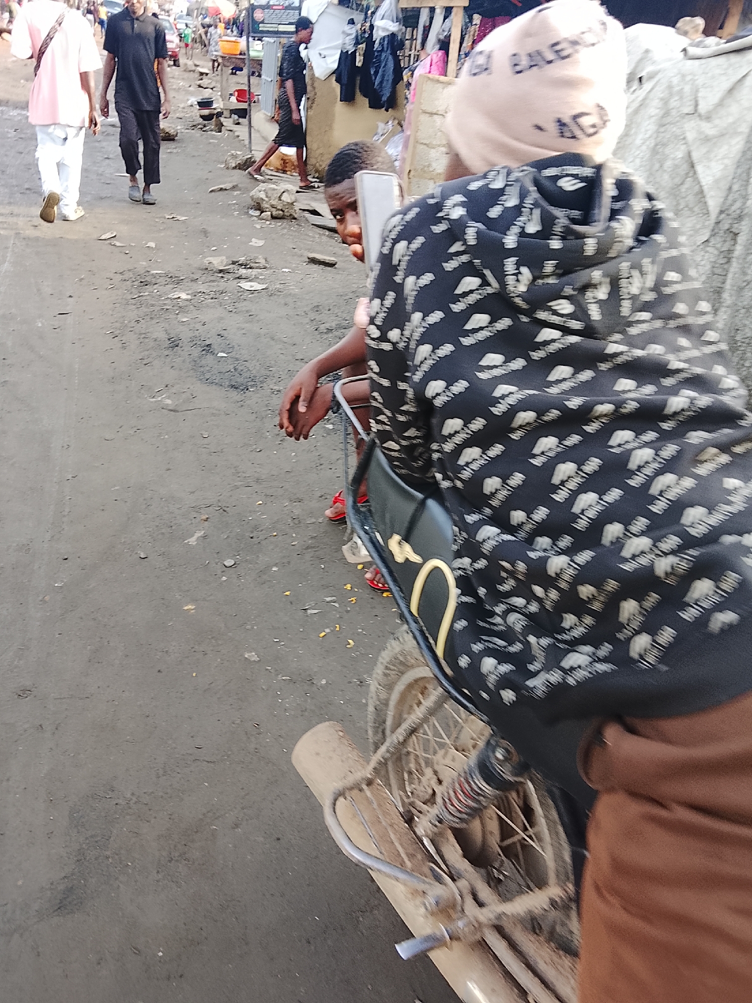 The image shows a dusty street scene in Abuja, Nigeria. In the foreground, a person wearing a Balenciaga-branded beanie and a black hoodie with the same branding is riding a motorcycle, partially obscured by the rider. Another person peers out from behind the motorcycle. In the background, people are walking on the street, some in Western-style clothes. There are buildings and stalls lining the street. The street is unpaved and dusty.