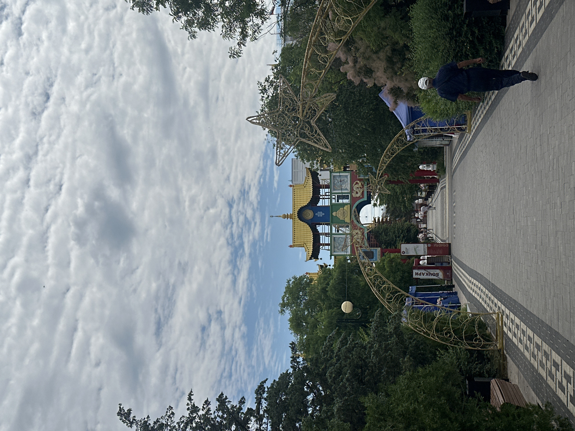 The image shows a person walking down a pedestrian walkway in Elista, Russia. The walkway is paved with gray stones and decorated with a black and white patterned border. On either side of the walkway are trees, bushes, and decorative gold arches with star motifs. In the background, there is a brightly colored building with a golden, multi-tiered roof in a distinct East Asian architectural style, characteristic of the city's Buddhist heritage. The sky is filled with white, fluffy clouds. Several stalls are set up along the walkway, with blue tents and signage. People are visible walking in the distance towards the building. The overall scene depicts a public area with cultural elements.