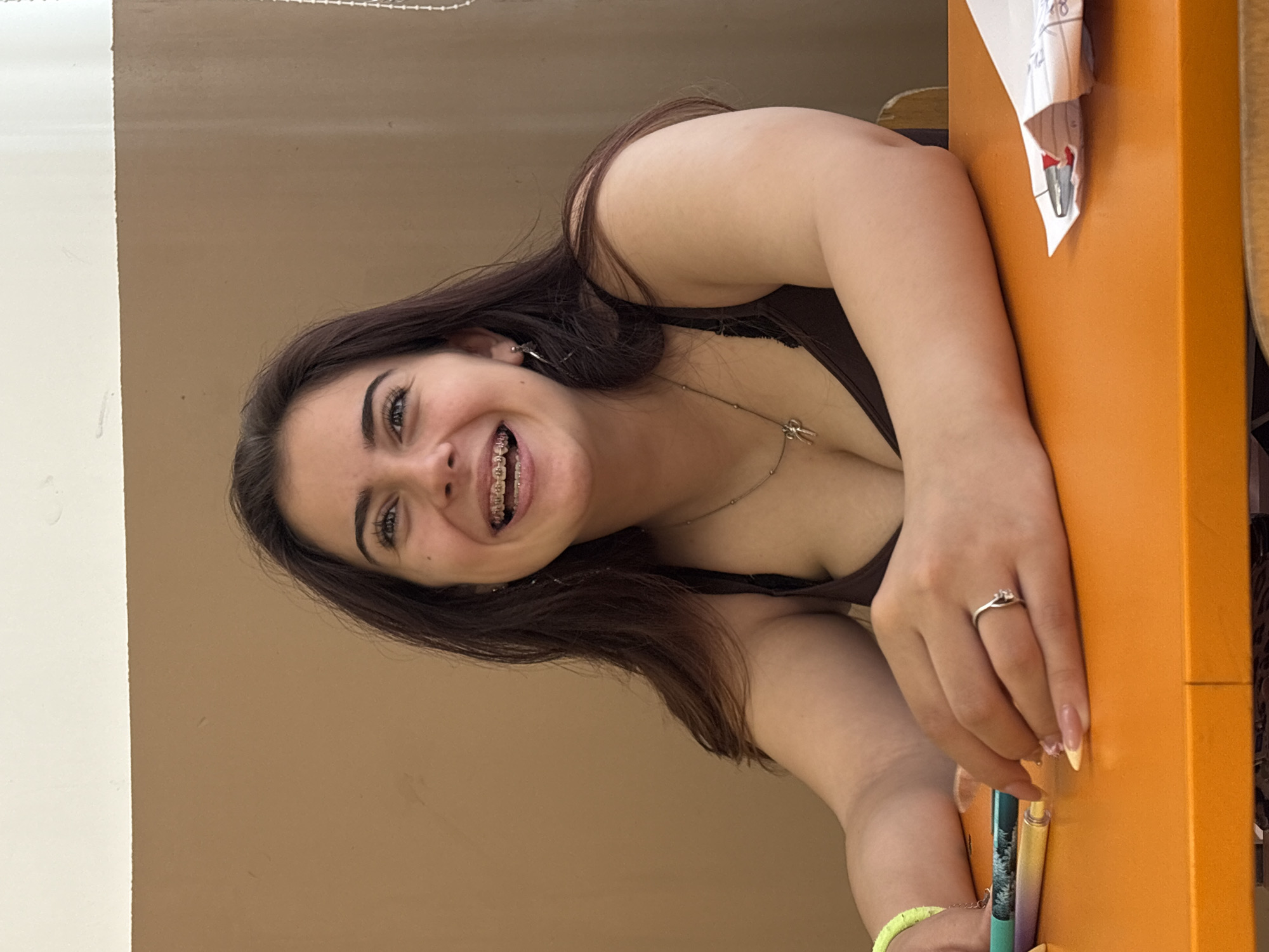 The image displays a young woman with long, dark hair and dark eyes, smiling cheerfully at the camera. She has braces on her teeth, which are visible as she smiles broadly. She is wearing a dark-colored top, possibly a tank top or camisole, and a silver necklace with a small charm around her neck. On her right hand, she wears a silver ring on her ring finger, and her nails are manicured with a light base and yellow tips. A bright yellow bracelet is visible on her left wrist.
She appears to be seated or leaning at an orange desk or table, with her right arm resting on the surface. Her gaze is directed at the viewer, and her expression is happy and friendly. Near her hand on the table are several writing instruments, including a teal/green pen and a yellow/gold pen. A crumpled piece of paper with some faint markings, potentially the number "28," is also present on the orange surface. The background behind her is a plain light brown wall, with a white wall or vertical surface on the left side of the frame. The setting appears to be indoors, suggesting a casual environment.