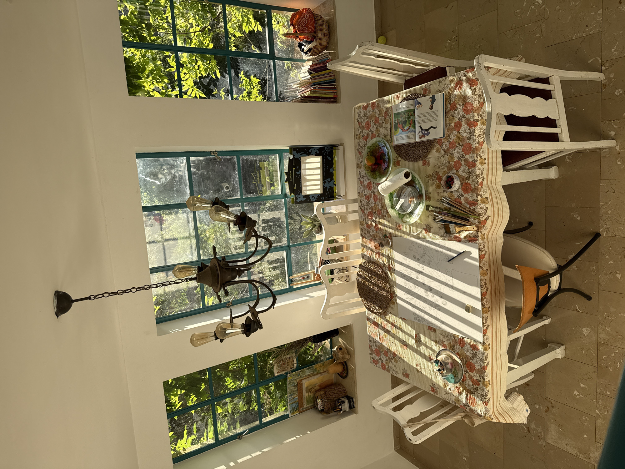 This image captures a brightly illuminated room, identified as being in Kfar Vradim, Israel. The room, likely a dining or activity area, is centered around a rectangular table with a floral tablecloth featuring patterns of red, orange, green, and cream.
On the table, there is a large white sheet of paper displaying a pencil drawing of what appears to be a vase with a plant or floral arrangement. Adjacent to the drawing are various art supplies including a cup holding paintbrushes, small containers of pigments, and an eraser. Other items on the table include a small woven placemat, a stack of books or a magazine opened to an illustrated page, a glass plate holding assorted fruits (citrus and red berries), and another glass plate with a roll of paper towel and a clear container of small items.
Surrounding the table are five white, painted wooden chairs with curved backs; one features a dark red cushion. An additional small chair with an orange seat and a black metal frame is partially visible tucked under the table on the left side.
Three distinct windows with dark green frames are set into the white walls, revealing lush green foliage outside. The leftmost window is angled inward at the top, creating a triangular section of wall above it. Strong sunlight streams through these windows, casting prominent, parallel shadows from the window frames across the table and the light-colored tiled floor.
A rustic, dark-toned metal chandelier with exposed Edison-style light bulbs hangs from the white ceiling, centered above the table.
The window sills display various objects. The left sill holds framed artwork, a wicker basket containing items, a gourd-like decorative object, and a black container with writing implements. The middle sill features a black decorative box with cut-out shapes (possibly a miniature stage or light box) and a small potted green plant. The rightmost sill is lined with numerous upright books, a wicker basket filled with textiles (an orange bandana is visible), and another small potted plant. A yellow ball is visible on the floor near the rightmost chair.
