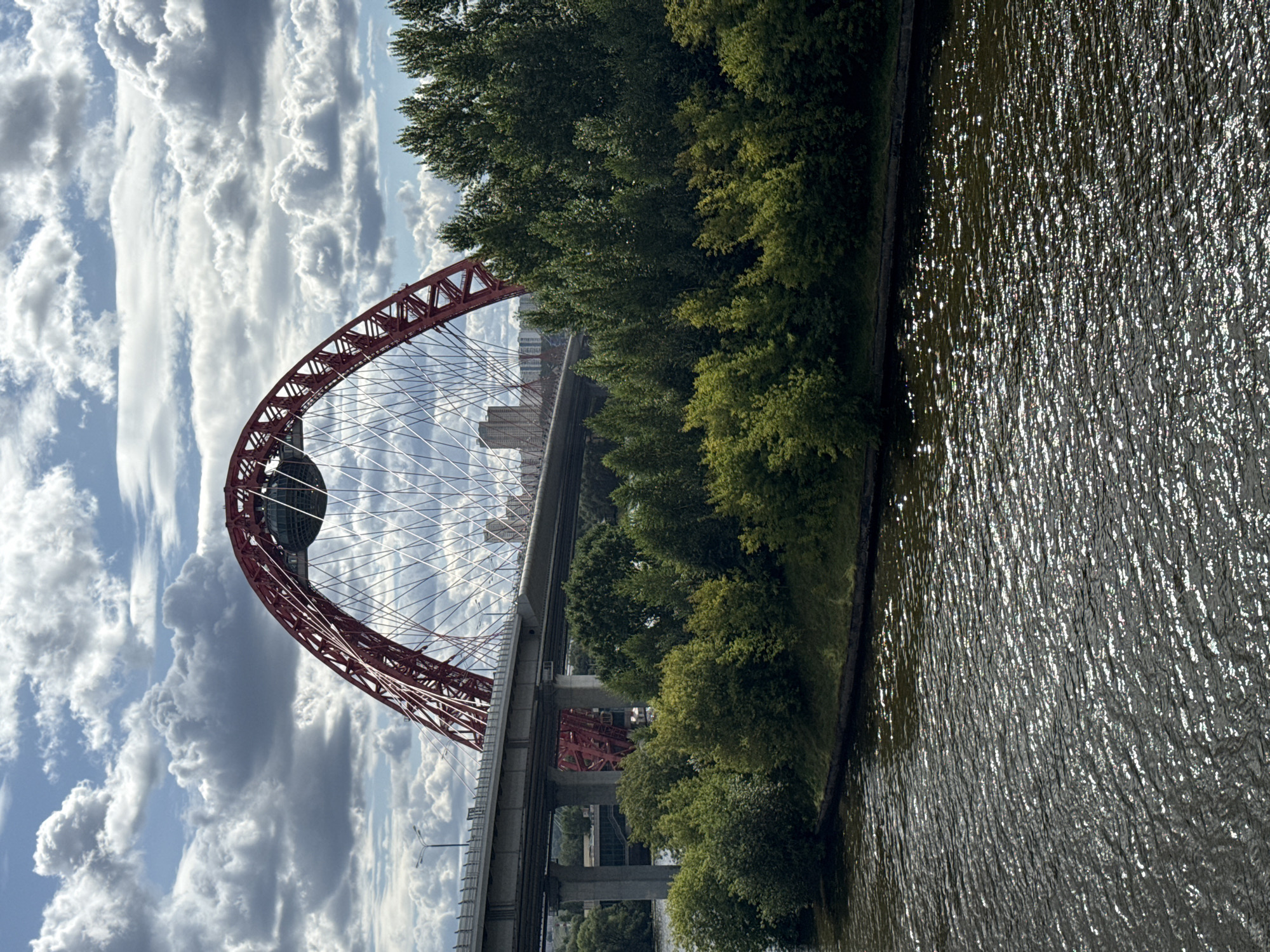 This is a photograph of the picturesque bridge, also known as the "Big Ice Cream" bridge, in Khimki, Russia. The iconic red arch of the bridge dominates the upper portion of the image, with its intricate network of cables stretching across the frame. Below the red arch, a second, gray bridge with concrete pillars is visible, suggesting a multi-layered transportation infrastructure. The foreground is occupied by a body of water, likely a river or canal, with its surface reflecting the dappled sunlight. Along the embankment on the right side of the image, lush green trees and vegetation form a dense line, partially obscuring the view of the city skyline in the background, where several tall buildings can be seen. The sky above is a mix of blue sky and dramatic, fluffy white clouds, adding depth and texture to the scene. The overall impression is one of a modern urban landscape integrated with natural elements.