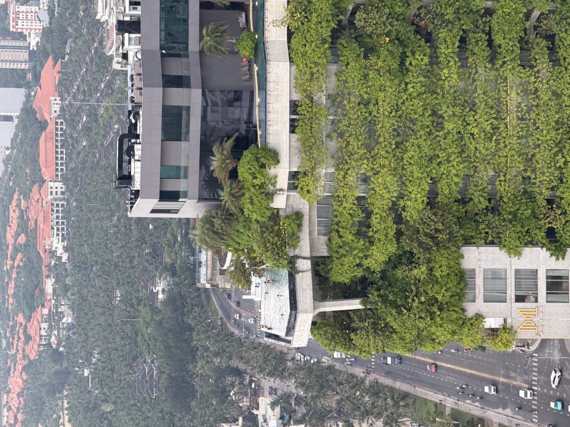 This is an aerial photograph taken in Đà Nẵng, Vietnam, with the image rotated 90 degrees counterclockwise. The scene depicts a modern hotel building with extensive greenery cascading down its facade.
The building features a prominent, elongated swimming pool on an elevated level, with several people swimming and relaxing. Palm trees are scattered around the pool area and along the surrounding streets. Adjacent to the hotel, a busy road is visible with cars and motorcycles navigating the lanes. Further away, rows of buildings with red-tiled roofs and other structures are seen, interspersed with dense palm tree foliage.
The time of day appears to be late afternoon or early evening, as suggested by the soft lighting and shadows. The weather seems pleasant, with clear skies. The overall impression is one of a luxurious urban resort environment, blending modern architecture with lush tropical vegetation.
Visible text includes "HOTEL" on the side of the building in large yellow letters.