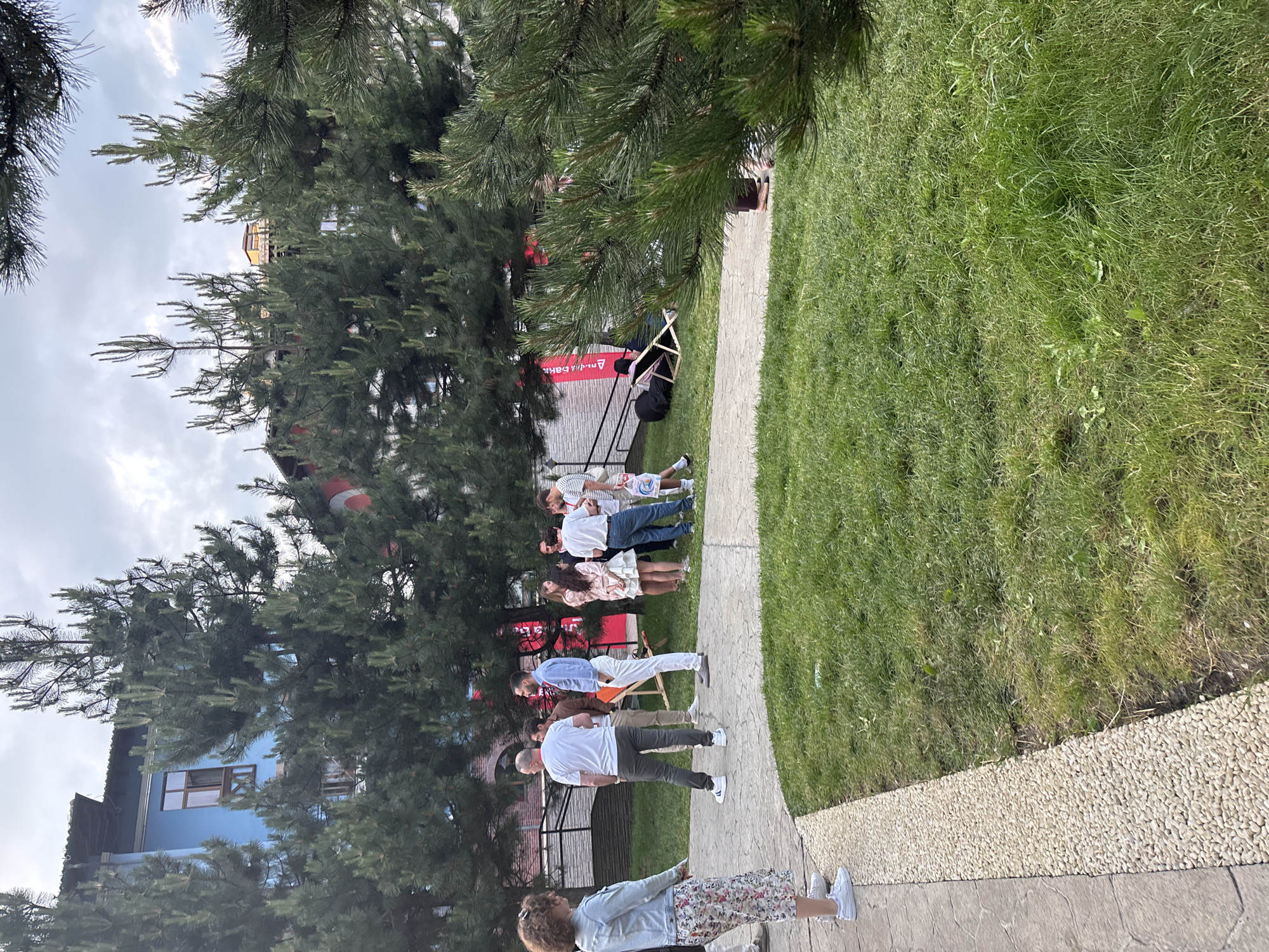 The image depicts a group of people gathered outdoors in what appears to be a park or a backyard. The setting features lush green grass, a paved walkway, and mature pine trees providing shade. Several individuals are standing in a line along the pathway, facing away from the camera. They are dressed in casual attire, suggesting a relaxed social gathering or event.
One person in the foreground, a woman with curly hair, wears a patterned skirt and a light-colored jacket. Behind her, a man in a white shirt and dark pants is also standing. Further back, a line of people in white shirts and various colored pants are visible, some appearing to be engaged in an activity. In the background, a blue building with wooden window frames can be seen peeking through the trees, and a red banner with white text is also visible.
The time of day seems to be daytime, likely late afternoon or early evening, given the quality of light and the presence of shadows. The weather appears to be pleasant, with the sun casting light on the grass, though the sky is partly cloudy. The overall mood of the scene seems calm and convivial. The visible text on the red banner is "АЛЬФА-БАНК", which is a Russian bank. The context of "Estosadok, Russia" suggests this location is in the Krasnodar Krai region of Russia, known for its mountain resorts.