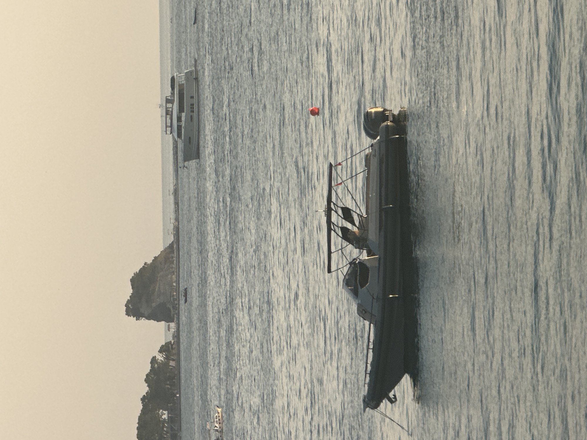 A motorboat is anchored in the water off the coast of Budva, Montenegro. The boat is dark gray with a canopy. In the background, a larger yacht is moored further out to sea. A small, rocky island with trees is visible to the left, and a red buoy floats in the water near the motorboat. The sky is a pale orange, suggesting either sunrise or sunset.