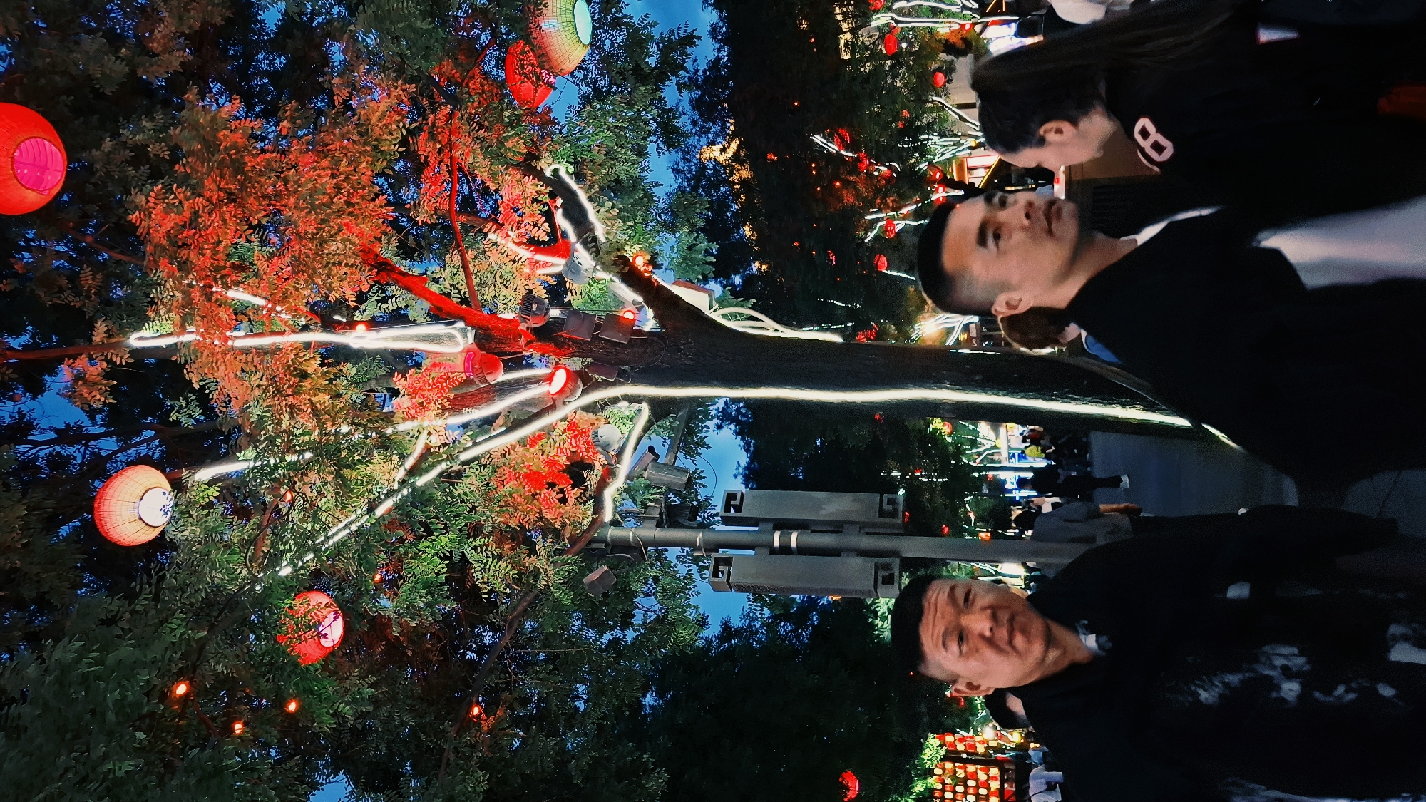The image depicts a street scene at night, likely in Yanta District, China, based on the presence of Chinese lanterns and architectural elements. The foreground features two men looking at the camera, with a woman positioned behind one of them. They are all wearing casual clothing. In the background, a tree is adorned with red Chinese lanterns and white string lights. Other people are visible walking on the sidewalk. There are also signs and streetlights visible.