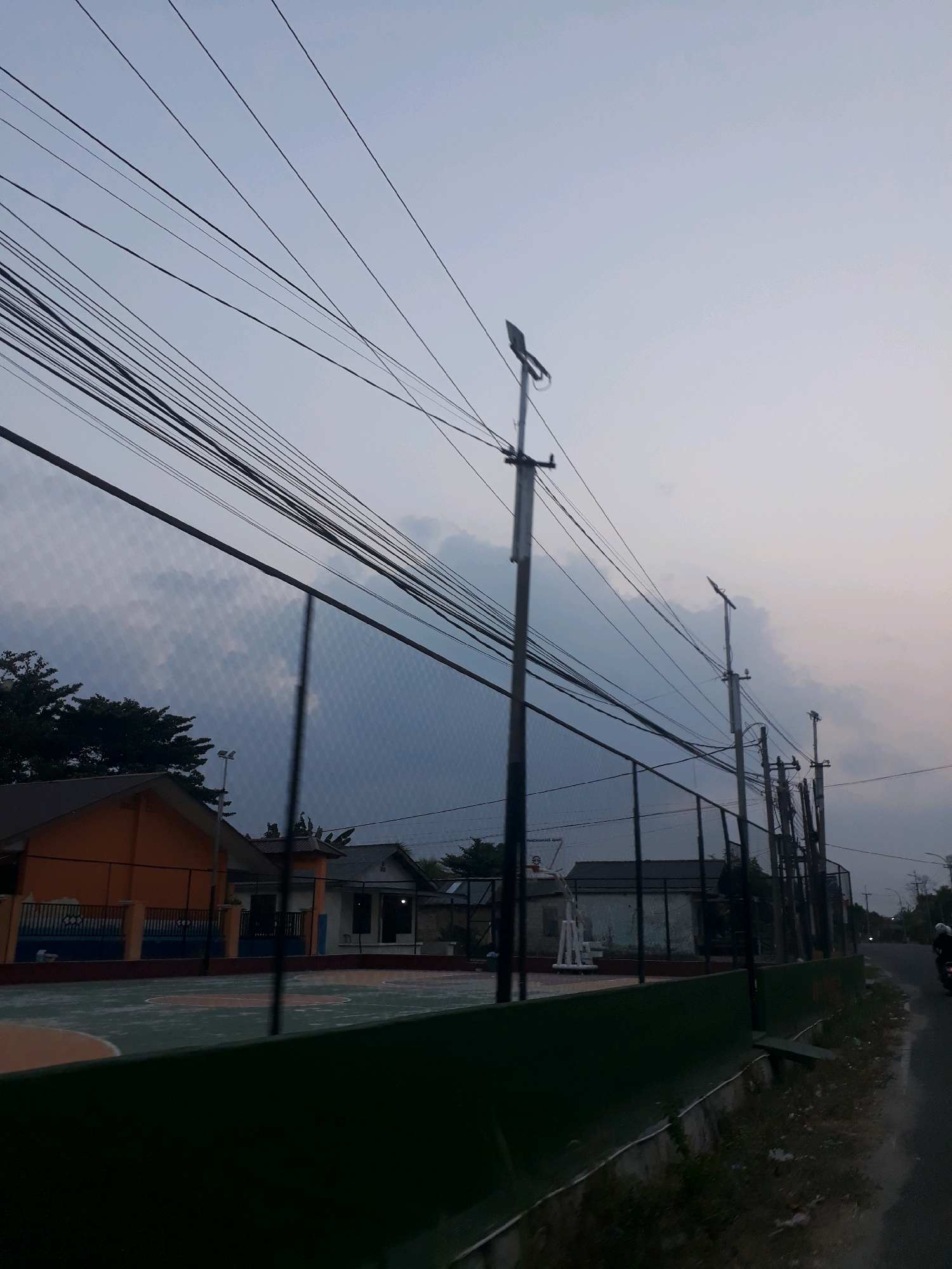 The image depicts an outdoor basketball court in the city of Pangkalpinang, Indonesia, during what appears to be dusk. The court is enclosed by a fence, and in the background, there are buildings and trees. Several utility poles with a dense network of wires are visible, stretching across the frame against a pale sky with some cloud cover. The lighting suggests it is either early morning or late evening. The court itself has markings, and a basketball hoop structure is present. To the right of the court, a road runs alongside, and a motorcycle with a rider is partially visible. The overall scene suggests a public or community sports facility.