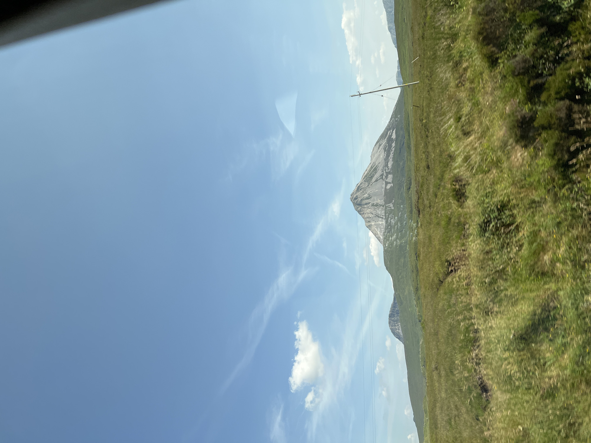 A daytime, eye-level, outdoor shot captures a panoramic view of a mountainous landscape under a bright blue sky dotted with wispy white clouds. The dominant feature is a large, grey, pointed mountain with visible lighter-colored striations, centrally located in the mid-ground. To its left, a smaller, rounded, dark grey mountain is visible. The foreground is a lush, green, undulating field of tall grass, with some areas appearing dry or brown, suggesting a natural, uncultivated environment. A single wooden telephone pole stands on the right side of the frame, angled towards the right, with thin wires extending horizontally across the scene. The overall impression is one of a rural, undeveloped, and possibly remote location. The scene is likely captured from a moving vehicle, as indicated by the slight blur in the foreground grass. The location is Meenagall, Ireland.