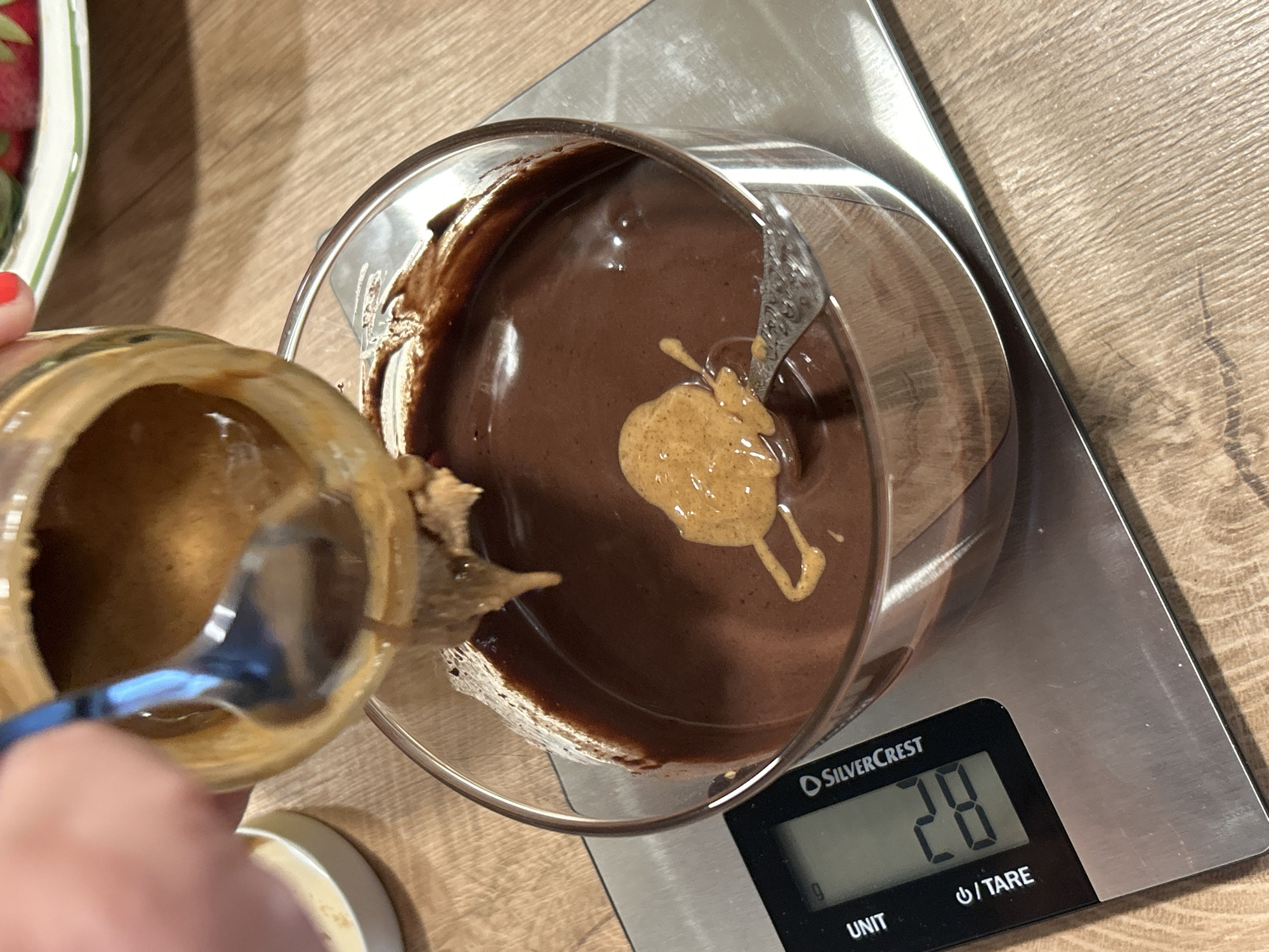 A hand is using a spoon to add peanut butter from a jar to a bowl of melted chocolate on a silver kitchen scale. The scale's display reads "28". The scene is indoors, set on a wooden-patterned surface.