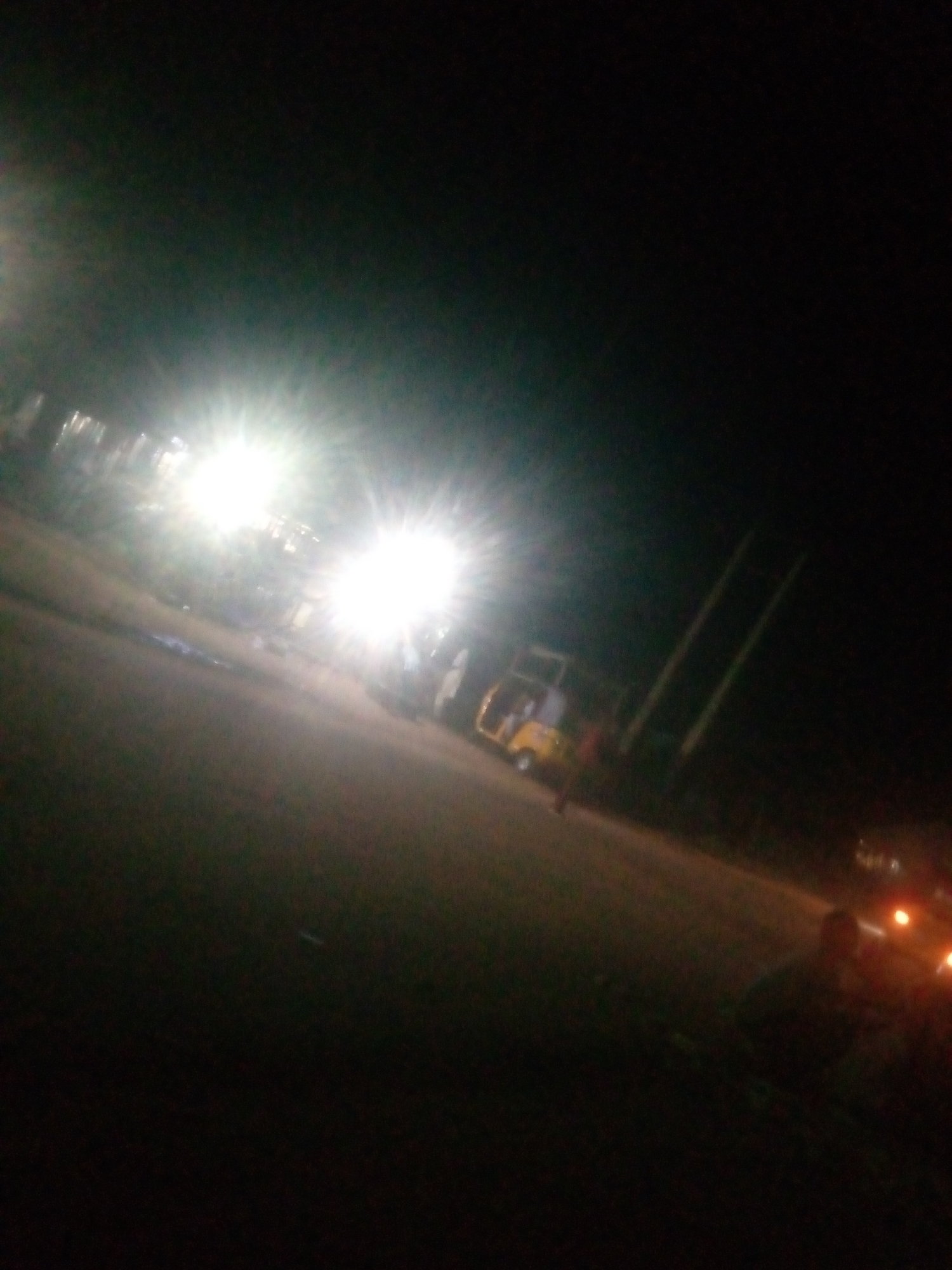 This is a blurry, low-light photograph taken at night in Jalingo, Nigeria. The scene appears to be a street or roadside with a few vehicles. Two very bright lights, likely from a vehicle or streetlights, are prominent, causing lens flare and obscuring details in their immediate vicinity.
In the mid-ground, a yellow autorickshaw (also known as a "keke napep") is visible. It appears to be parked or moving slowly. A person, possibly the driver or a passenger, is dimly visible near the autorickshaw. Further in the distance, another vehicle with illuminated headlights is faintly discernible.
The overall atmosphere is dark, typical of nighttime in an area with limited street lighting. The ground appears to be a paved surface, possibly a road. There is no visible text in the image. The blurriness of the image suggests it may have been taken with a moving camera or in poor lighting conditions.