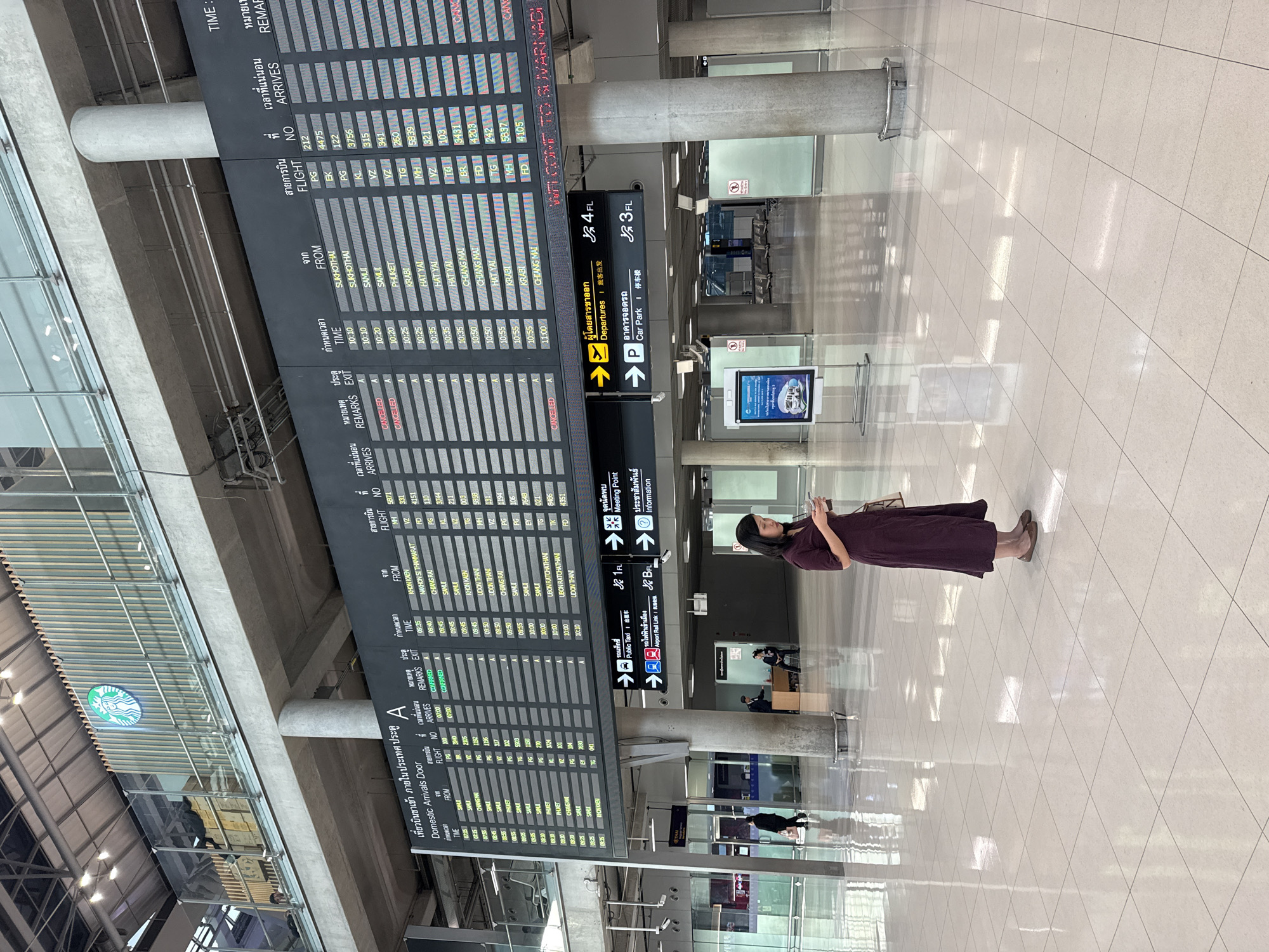 The image depicts the interior of a modern airport terminal, likely in the city of Nong Prue Subdistrict, Thailand, as evidenced by signage and the prominent "WELCOME TO SUVARNABHUMI" text. A large digital flight information board dominates the center of the frame, displaying flight arrivals and departures. The board is multi-lingual, featuring Thai and English text.
In the foreground, a woman in a dark purple dress stands looking at her phone, holding a drink in her right hand. She is positioned in a wide, brightly lit concourse with a polished white tile floor. Behind her, an information desk is staffed by two individuals, and a few other travelers are visible in the distance. Overhead, a Starbucks logo is visible on a raised platform. Signage indicates directions for "Departures," "Public Taxi," and "Car Park." The overall environment is clean, spacious, and functional, characteristic of a major international airport.