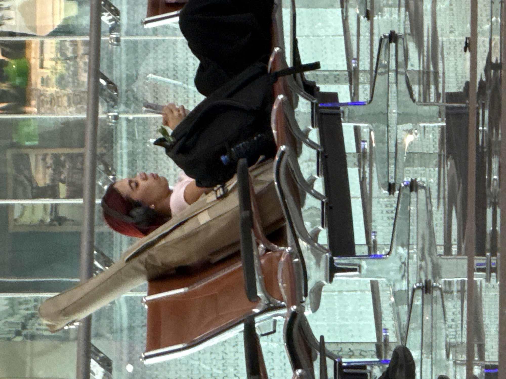The image shows a woman with red hair wearing headphones, sitting in an airport lounge in Seeb Province, Oman. She is holding a phone and looking at it, with a black backpack beside her and a tan case, possibly for a musical instrument, leaning against the back of her seat. The lounge has a modern design with reflective surfaces and rows of seating. The background appears to be a large window overlooking an indoor area with more seating and a view of a city skyline through glass.