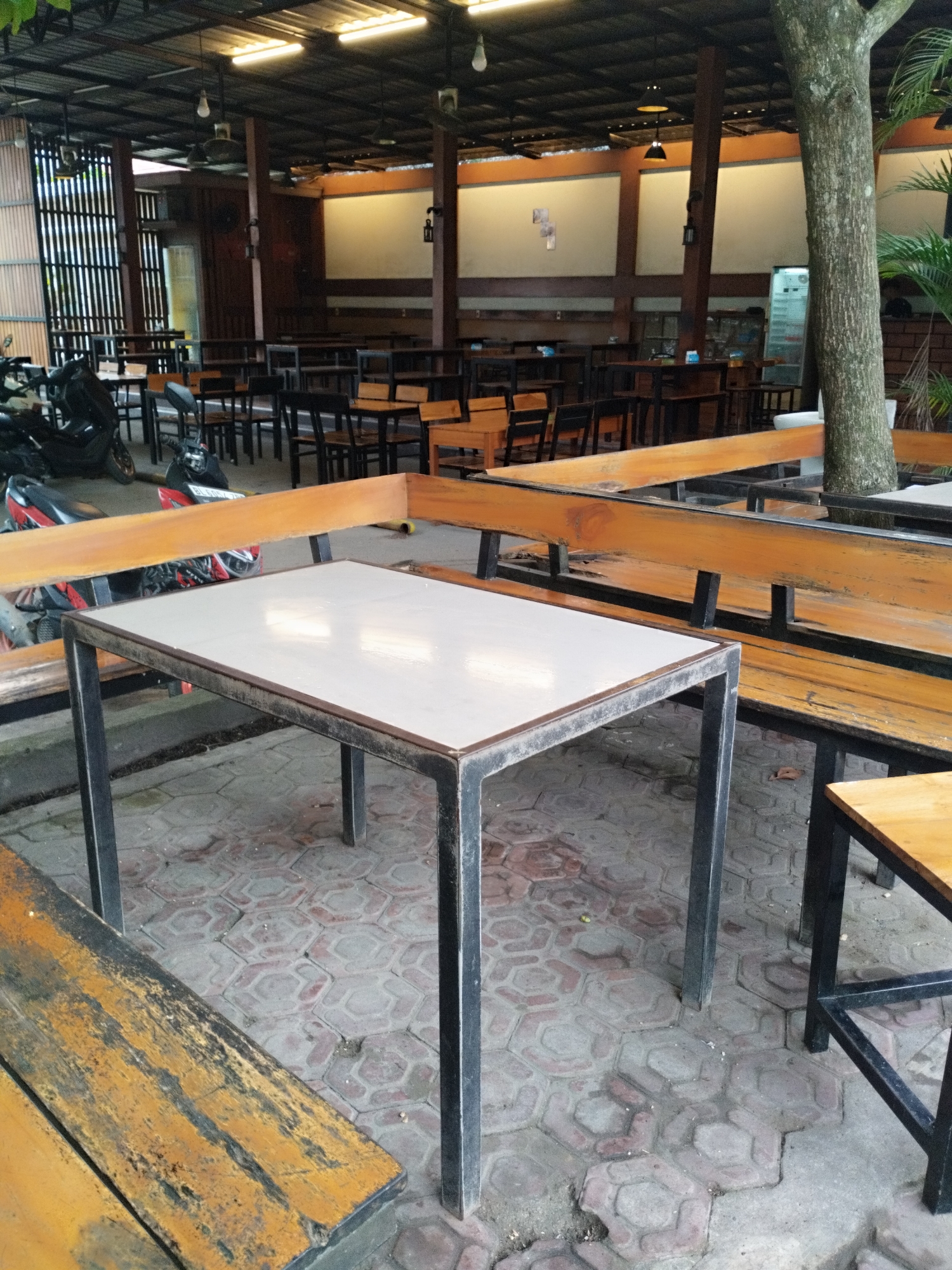 The image depicts an outdoor seating area of a restaurant or cafe, likely in Payameuneng, Indonesia, characterized by its open-air design with a metal roof supported by wooden beams. Several tables and benches are visible, constructed with a combination of wood and metal. In the foreground, a rectangular table with a white top and dark metal legs is prominently featured. To the left and right of this table are wooden benches with metal supports. The ground is paved with hexagonal tiles. In the background, more tables and chairs are arranged, some with wooden tops and dark metal frames. Motorcycles are parked to the left of the seating area, suggesting it might be a common mode of transportation for patrons. A large tree trunk is visible on the right side of the image, indicating a natural element integrated into the outdoor space. The overall impression is one of a functional and somewhat rustic dining environment.