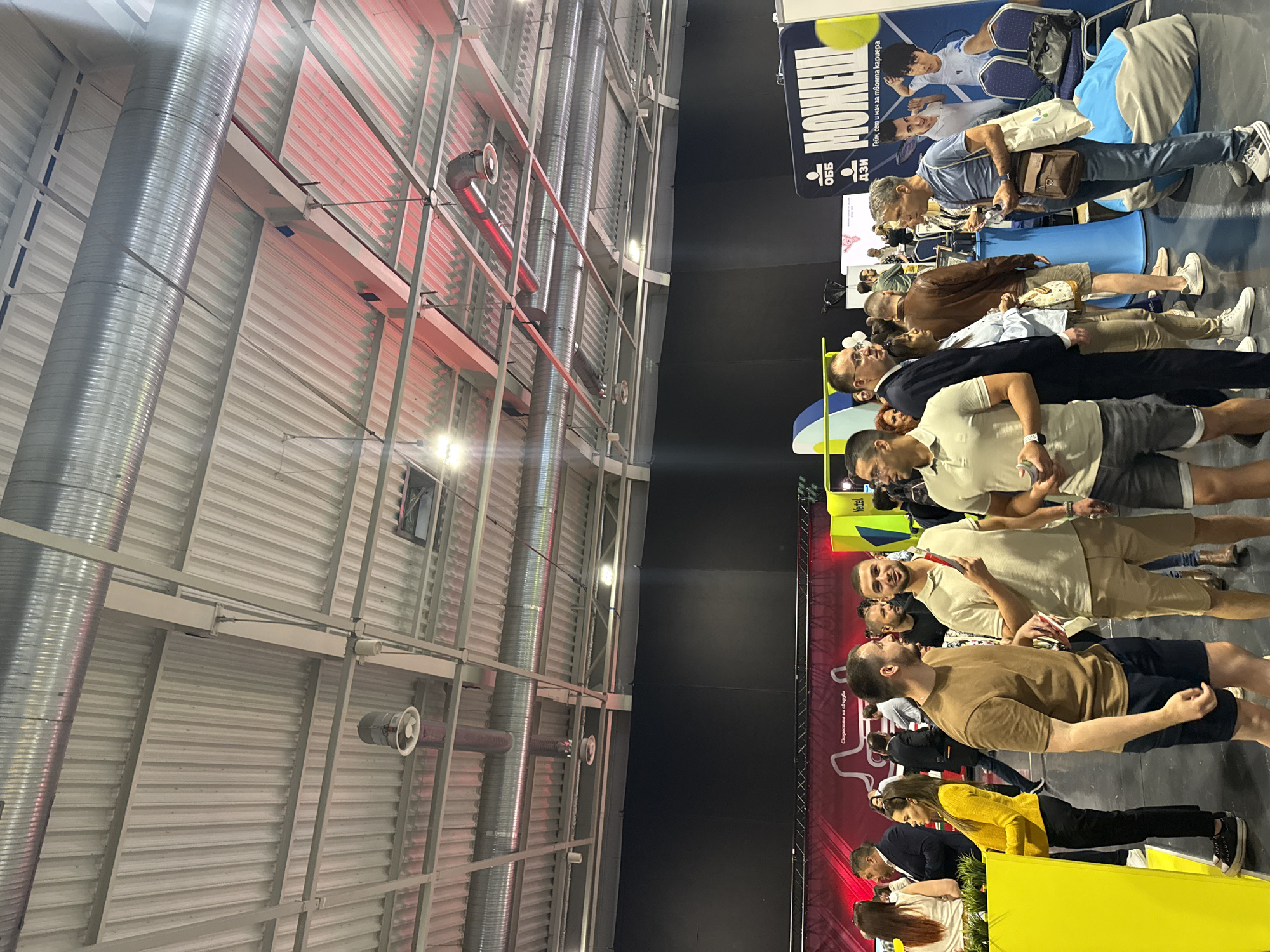 The image depicts a large indoor space in Sofia, Bulgaria, possibly an exhibition hall or convention center. A crowd of people stand in a line, primarily facing toward the right side of the frame. The individuals are dressed casually, and appear to be of various ages and ethnicities. Some are holding items in their hands, possibly drinks or promotional materials.
Behind the line of people is a dark backdrop, possibly a booth or display area for an event. There is a yellow counter or table in the foreground, with some branding or signage on it.
The ceiling is high and industrial, with exposed metal beams, ventilation ducts, and light fixtures. The lighting is bright, illuminating the scene. A large banner or sign in a foreign language is visible in the upper right.