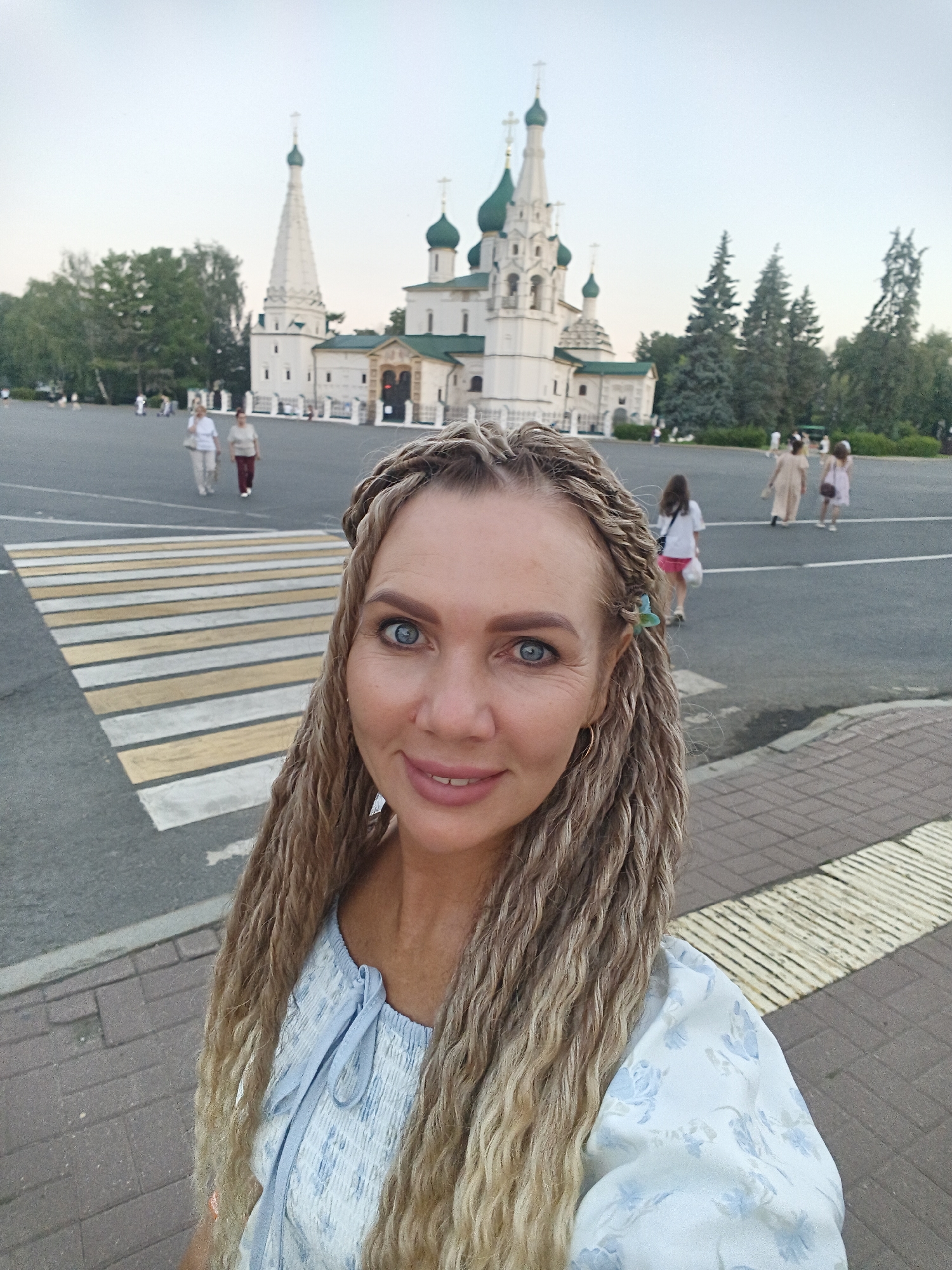 The image shows a woman in the foreground, taking a selfie. She has blonde hair styled in braids, blue eyes, and is wearing a light blue dress with a floral pattern. Behind her, a pedestrian crossing with yellow and white stripes is visible. In the background, a large white church with green domes and a tall steeple stands in a plaza. Several people are walking in the plaza, and trees are scattered throughout the area. The scene appears to be set in a public square in Yaroslavl, Russia, likely near the Assumption Cathedral. The sky is overcast, suggesting it might be late afternoon or early evening.