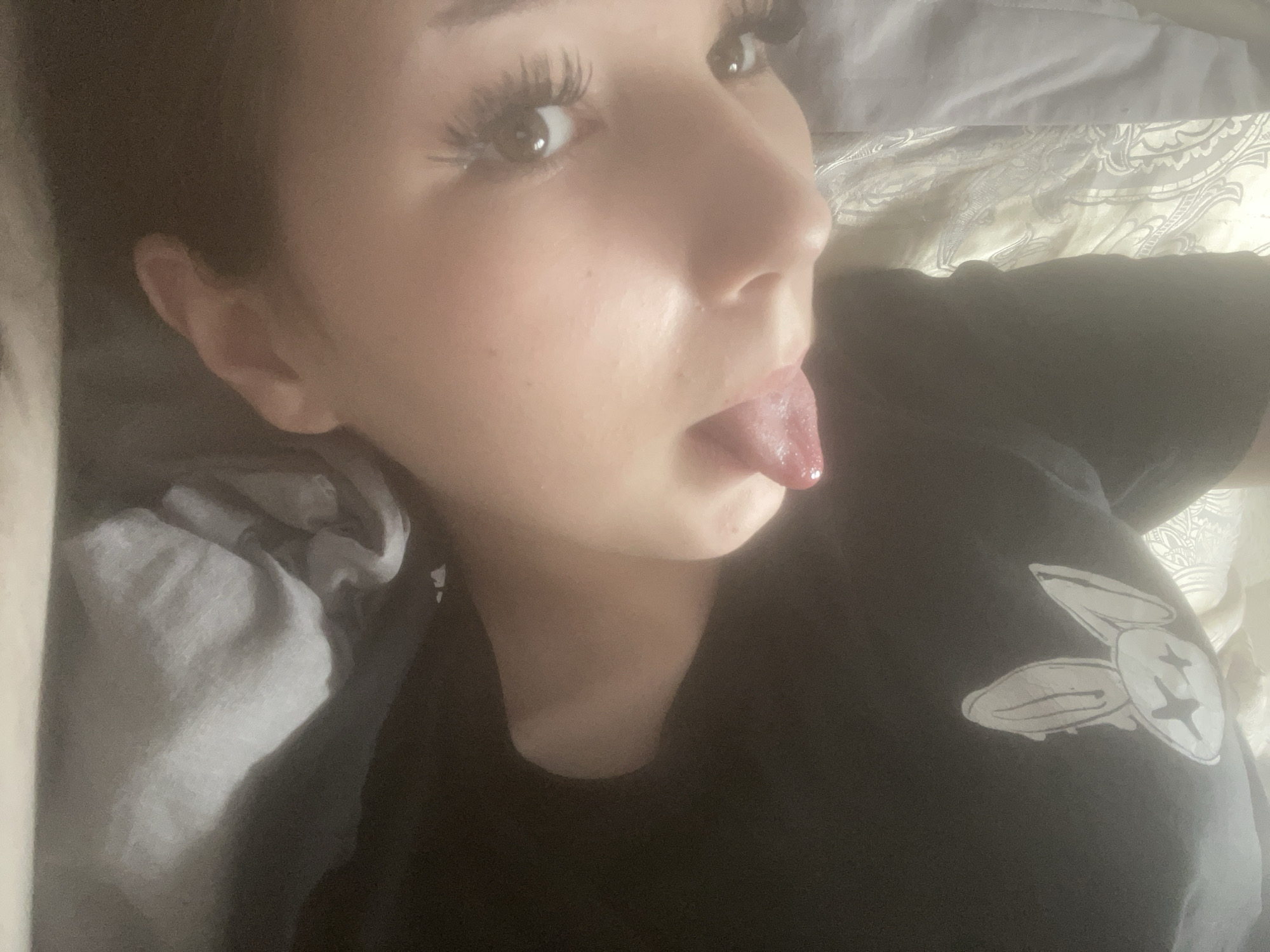 The image presents a close-up, angled portrait of a young person, likely female, lying down indoors, most probably in a bed. The subject's head is tilted, showing their right ear, part of their face including the right eye, nose, and mouth. They have long, dark eyelashes and a playful expression, with their tongue slightly extended.
The person is wearing a dark, possibly black, t-shirt or top. A distinct white graphic is visible on the chest area of the shirt. This graphic consists of two stylized shapes that resemble either cartoon rabbit ears or, more precisely, two flat-headed screws or bolts. One of the shapes clearly shows a cross-shaped indentation, consistent with a Phillips-head screw.
The background comprises crumpled bedding. On the left side of the frame, light grey or white fabric is visible, while on the right, a lighter-colored (beige or cream) sheet with a subtle patterned design, possibly paisley or damask, can be seen. The lighting in the image is soft and diffused, typical of an indoor setting.
The overall mood of the image is casual and lighthearted. There are no visible text elements in the media file, and no specific visual cues within the image itself denote the mentioned location of Sinyavino, Russia.