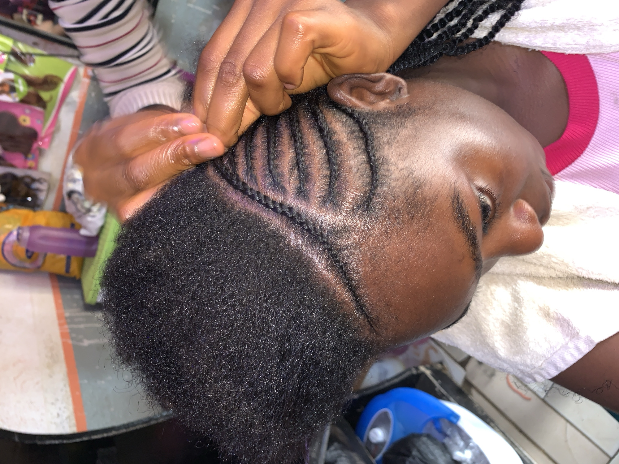 A person is getting their hair braided in a cornrow style. The person's head is tilted, and their eye is slightly visible. The braids are neat and tight, with a distinct pattern. Hands are seen working on the braids, appearing to be freshly oiled or moisturized. The background shows a cluttered environment with various items like hair products and containers. It is unclear where this scene is in Rafin Par, Nigeria.