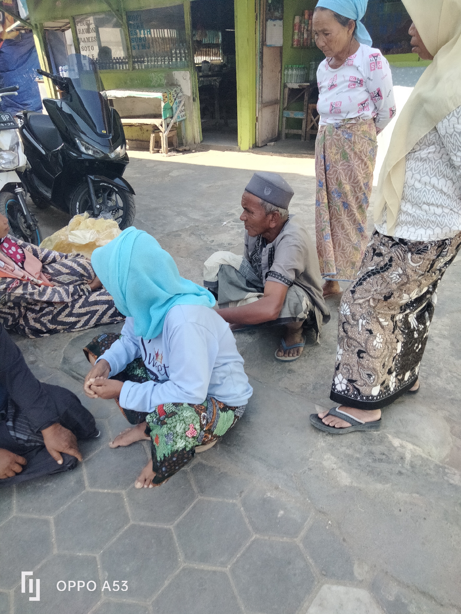 In the city of Badung, Indonesia, an outdoor scene unfolds on a paved ground with hexagonal tiles. A black motorcycle, parked in the foreground, is partially obscured by bags of goods. Several individuals are present. An older man, wearing a grey cap and shirt, squats on the ground. To his right, a woman in a light blue headscarf and patterned skirt is also seated. Another woman, partially visible, wears a patterned skirt and a light-colored headscarf. In the background, a green stall with a weathered sign displays various products. The overall atmosphere suggests a market or roadside setting.