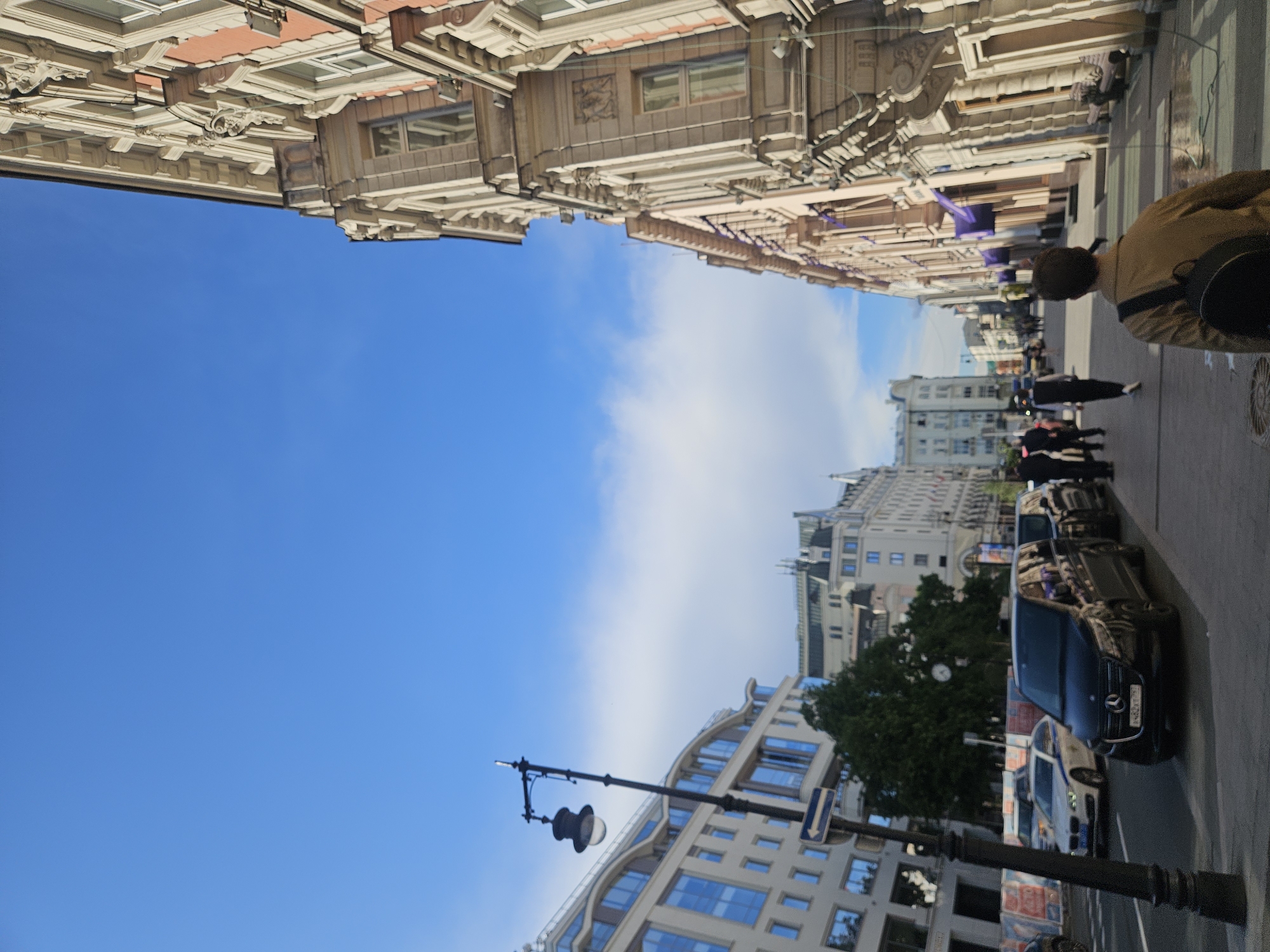 Here's a description of the image:
The image presents a street scene, likely taken in the city of Moscow, Russia. Buildings line both sides of the street, showcasing a variety of architectural styles. On the left side, there is a modern building with a curved facade and numerous windows. A lamp post is visible near the lower left corner. In the center, the sky is visible, with light clouds.
On the right side of the street, there are older, more ornate buildings. Several people are walking along the sidewalk. Parked cars are lined up on the side of the road, including a black car prominently featured. A person with a backpack is walking on the sidewalk, partially visible in the right side of the image. The scene appears to be set on a clear, sunny day.
