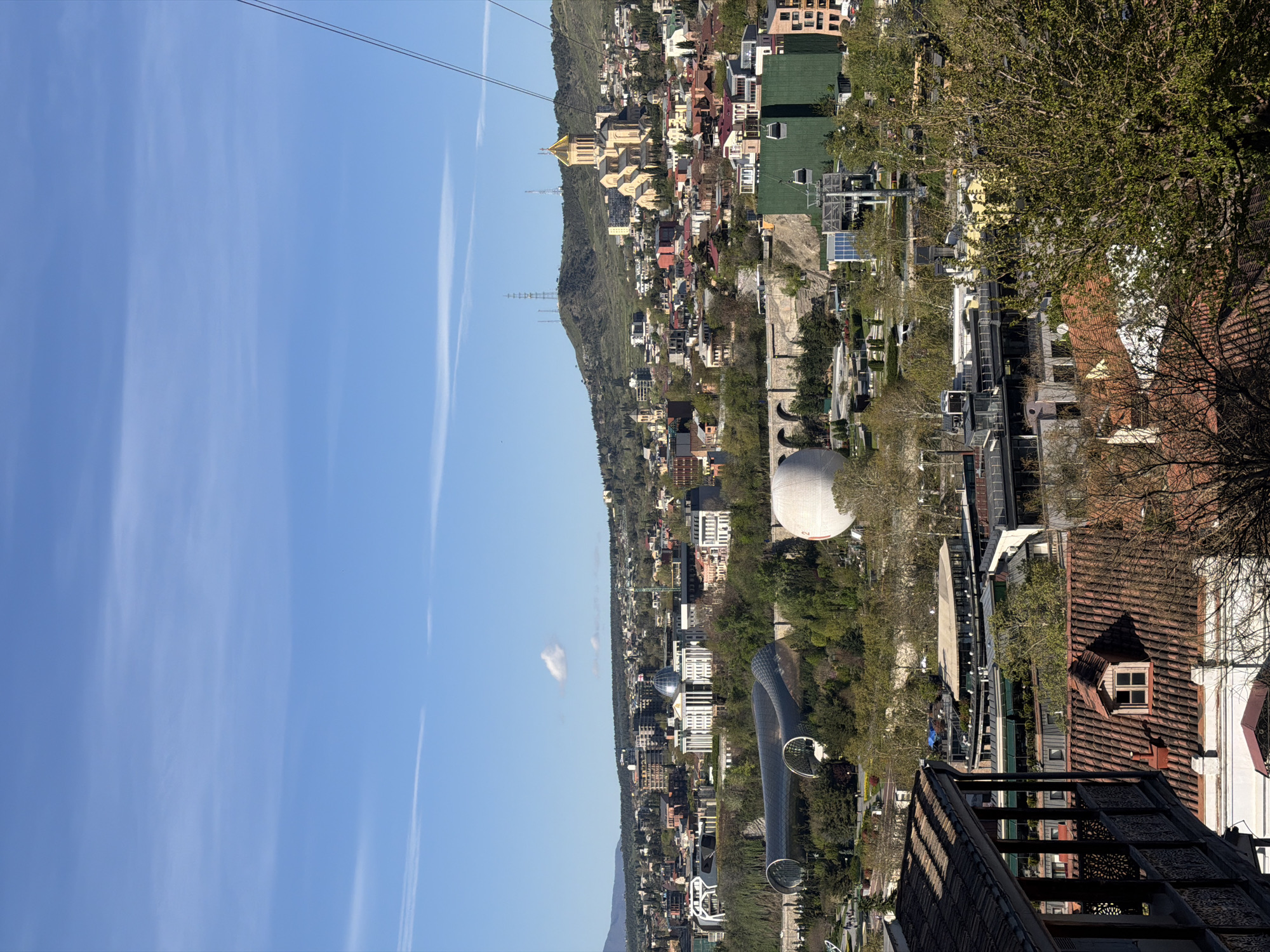 The image depicts an aerial view of Tbilisi, Georgia, shot from a high vantage point. In the foreground, part of a building with a brown tiled roof and a decorative metal structure is visible, suggesting the location is elevated. The cityscape includes a mix of residential and commercial buildings of varying heights and architectural styles. A large, white spherical structure is prominent, possibly a cultural or recreational facility. The Mtkvari River is not discernible in the image, but the cityscape is suggestive of the city's geography. Hills and mountains are visible in the background, contributing to the landscape. The sky is clear and blue with contrails visible. A cable car line runs across the top of the frame.
