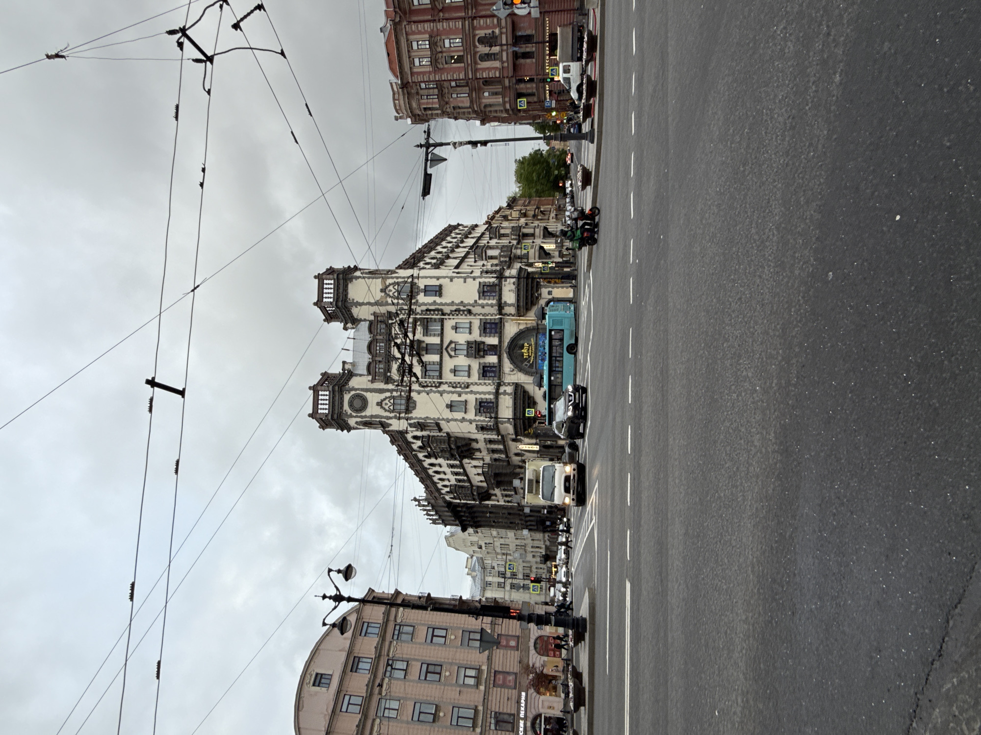 The image captures a street scene in Saint Petersburg, Russia, likely Nevsky Prospekt, distinguished by its architectural landmarks and urban setting. The street is paved with asphalt, marked by white lines indicating traffic lanes. A notable feature is a large, ornate building with a light-colored facade and clock towers, suggestive of early 20th-century architectural design. Other buildings line the street, showcasing a variety of architectural styles. Overhead, a network of wires spans across the sky. Traffic includes buses and other vehicles traveling along the street. Pedestrians are visible. The sky is overcast, casting a soft light over the scene.