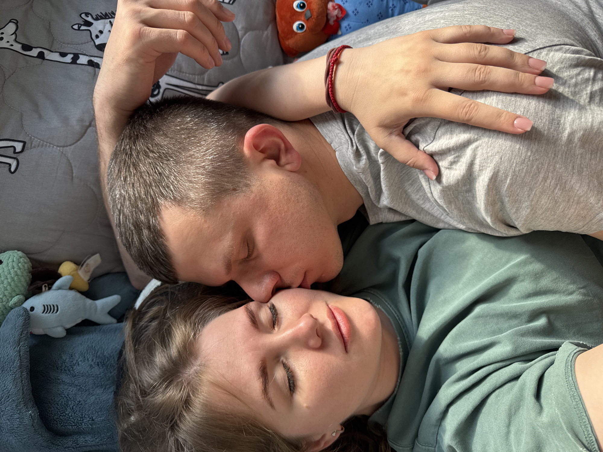 A high-angle, close-up shot shows a couple lying on a bed. A man with short, dark hair is kissing a woman's forehead. The woman has her eyes closed. He is wearing a gray t-shirt and a red bracelet. She is wearing a green t-shirt. Stuffed toys surround them on the bed, including a shark and an orange owl. The bedding has a pattern of cartoon animals. The location is most likely a private residence in Ферма, Russia.